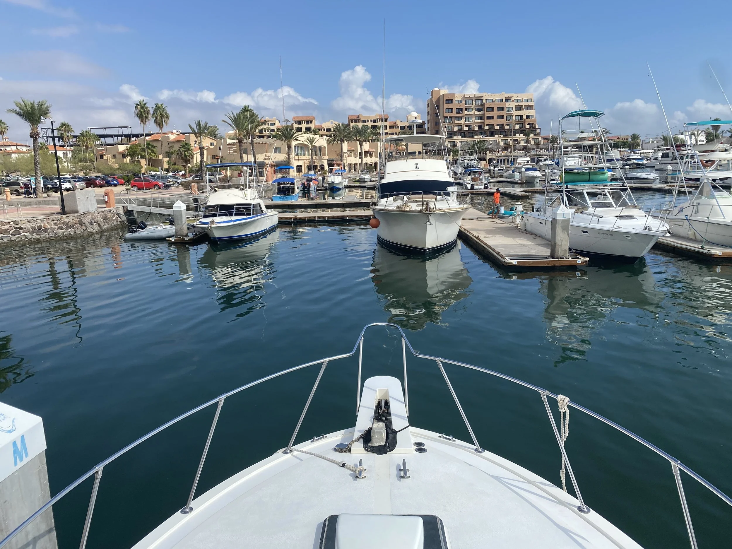 View from a boat looking toward a marina in San Carlos, Mexico with several docked boats, palm trees, parking lot, and buildings in the background on a clear day.