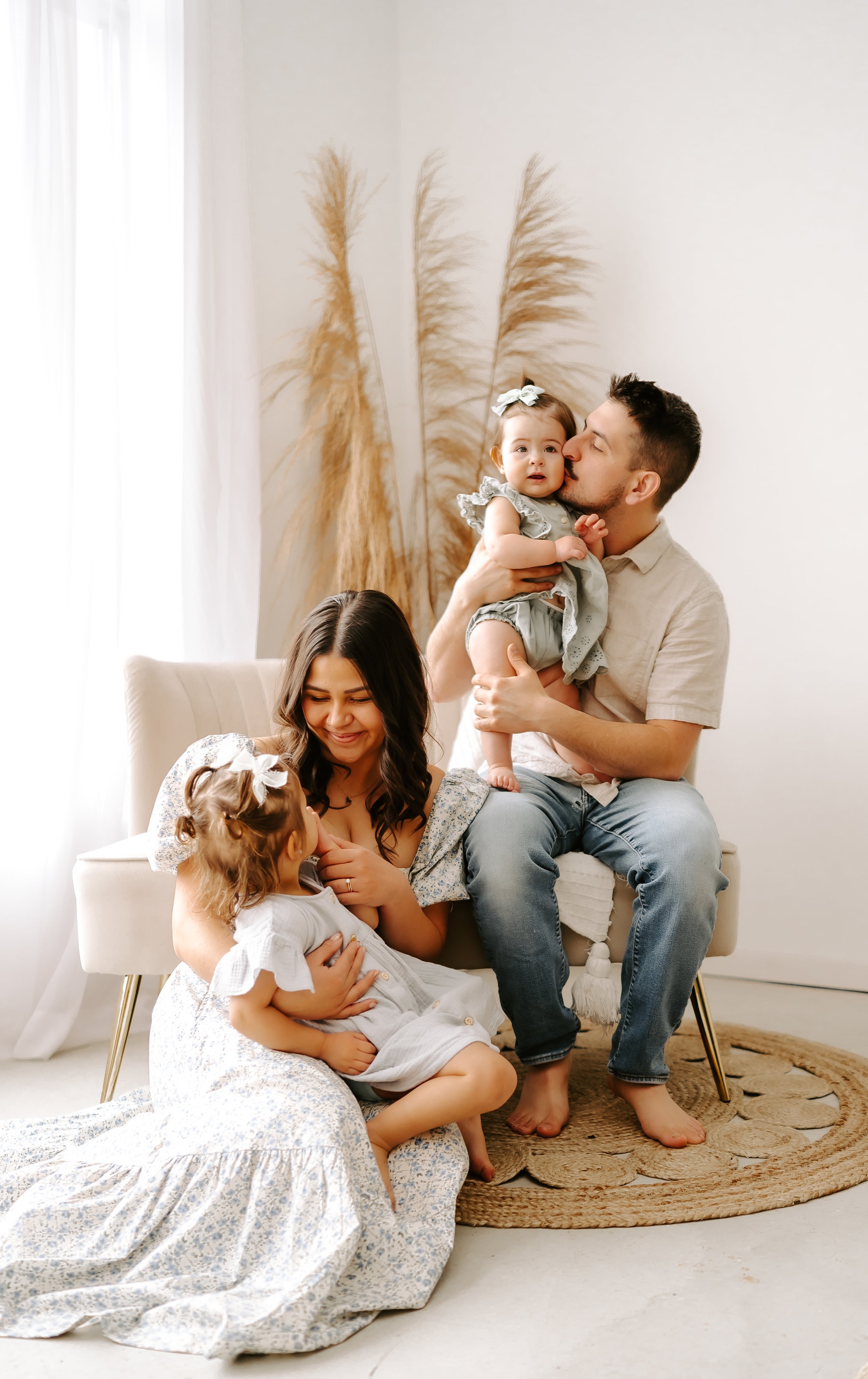 Family portrait with a man sitting on a chair holding a baby and a woman sitting on the floor with a toddler; pampas grass in the background.