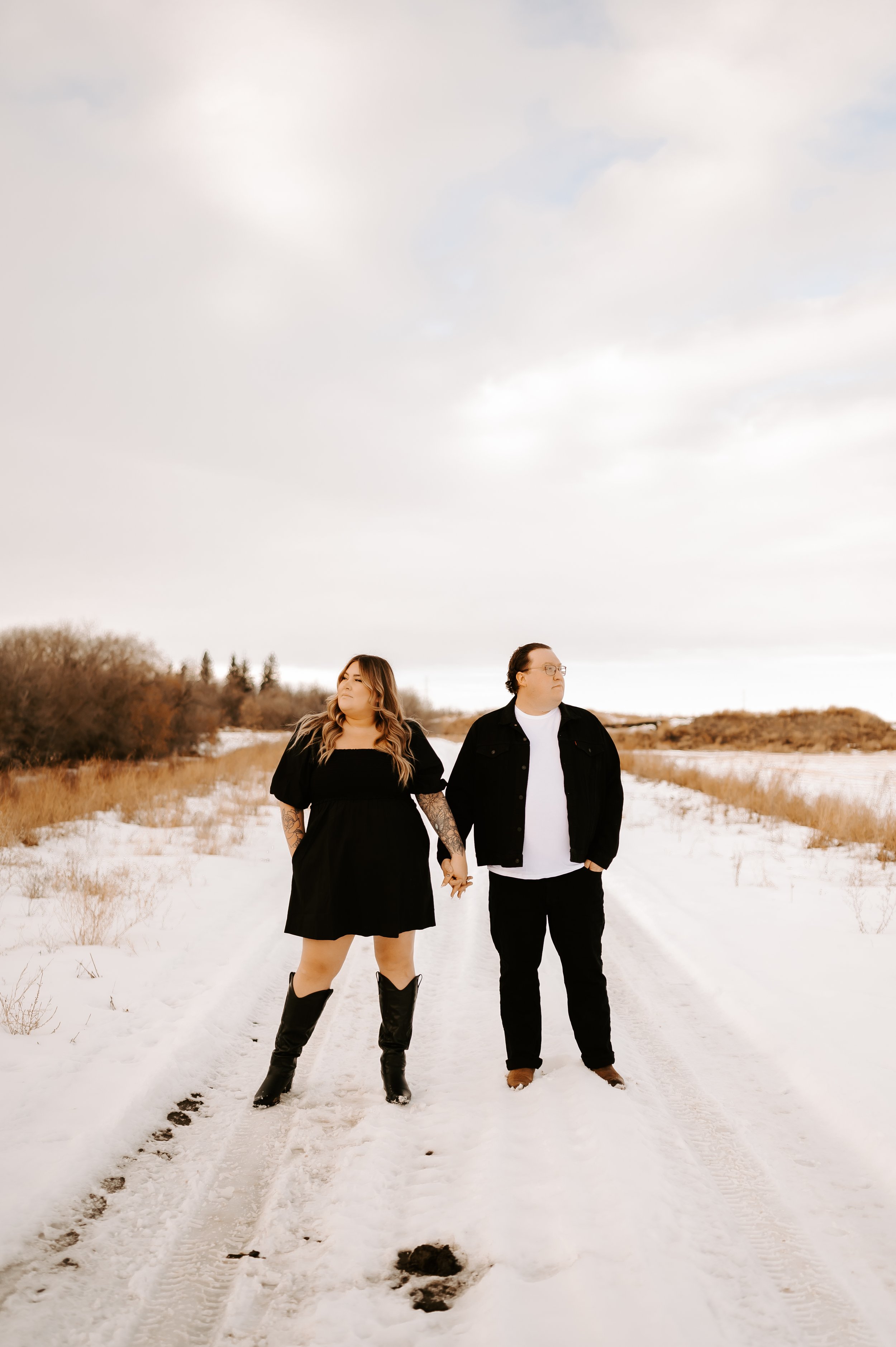 Couple holding hands standing on snowy road with cloudy sky.