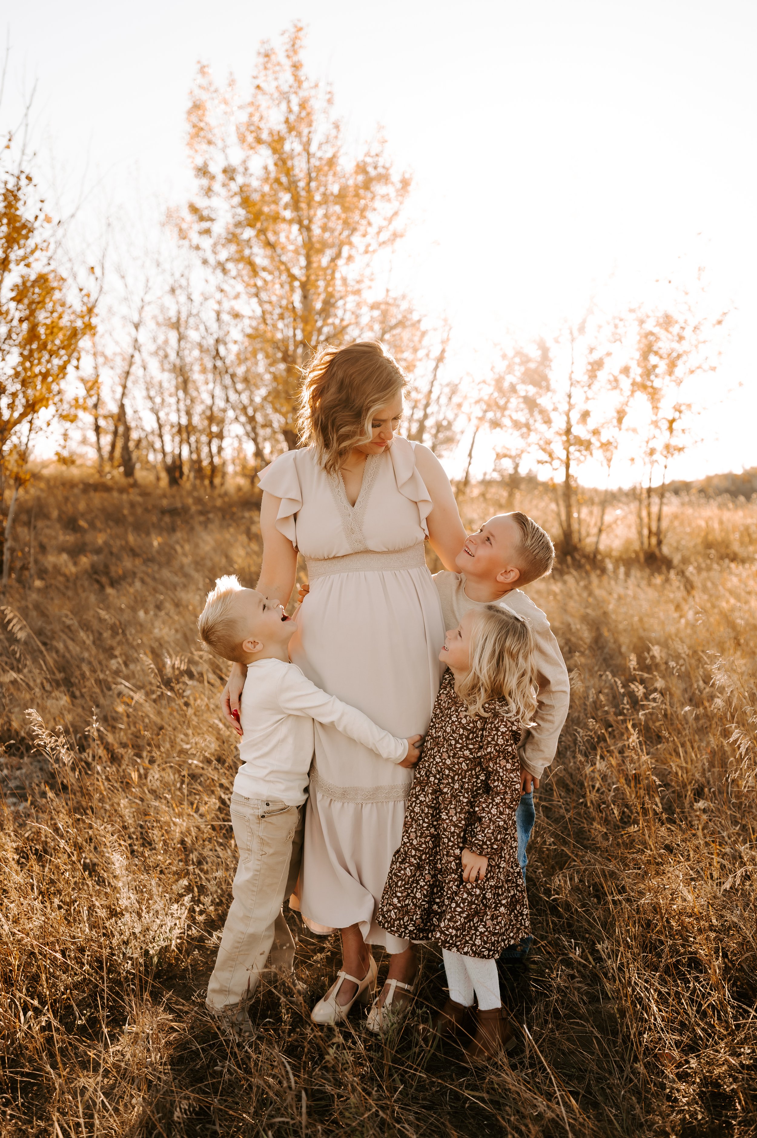 A woman in a light dress stands in a field with three children embracing her, surrounded by tall grass and autumn trees.