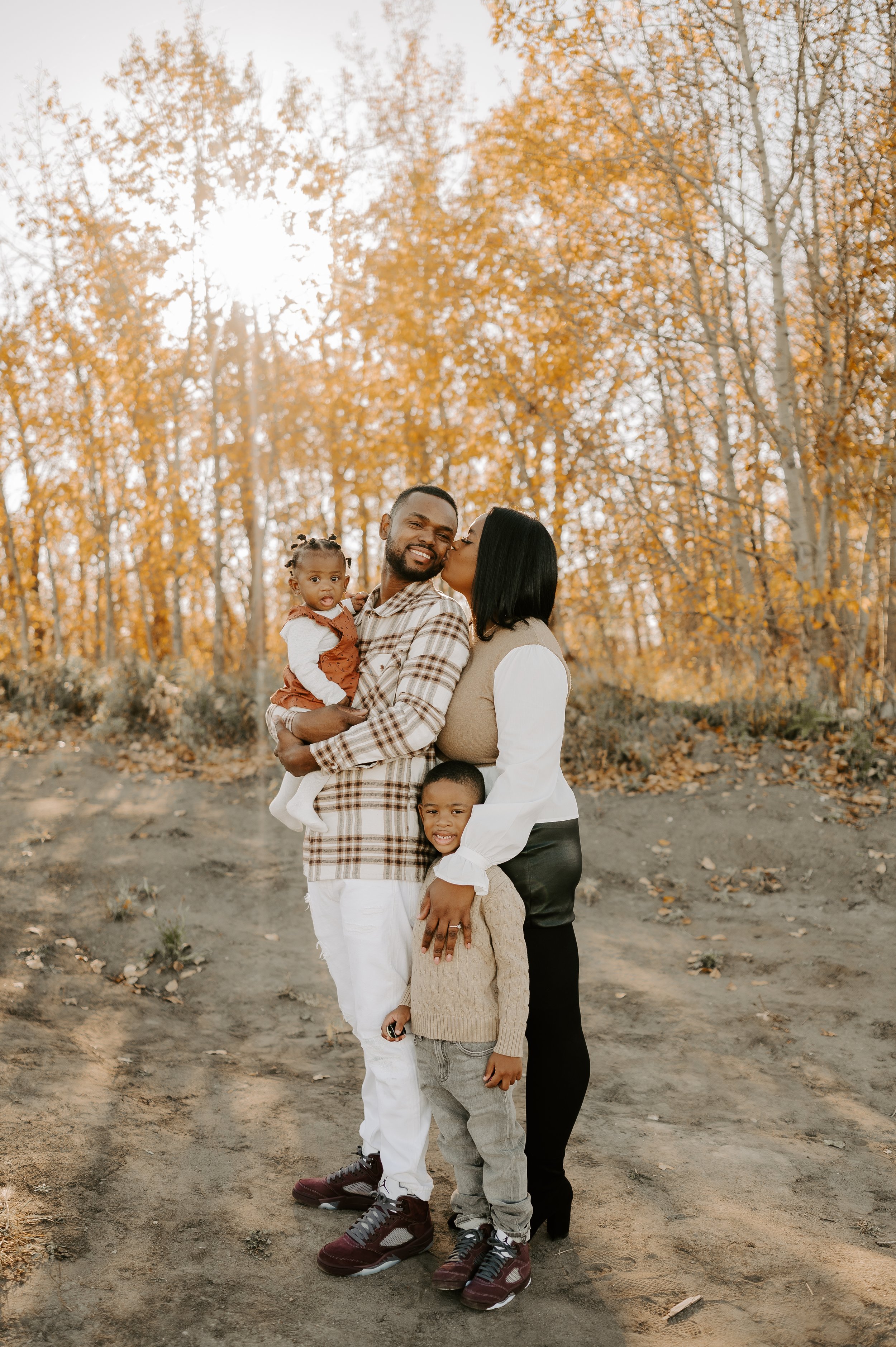 A family of four poses outdoors with fall foliage in the background, featuring golden trees. The parents are smiling, the mother kissing the father, while holding a baby. A young boy stands in front, smiling.