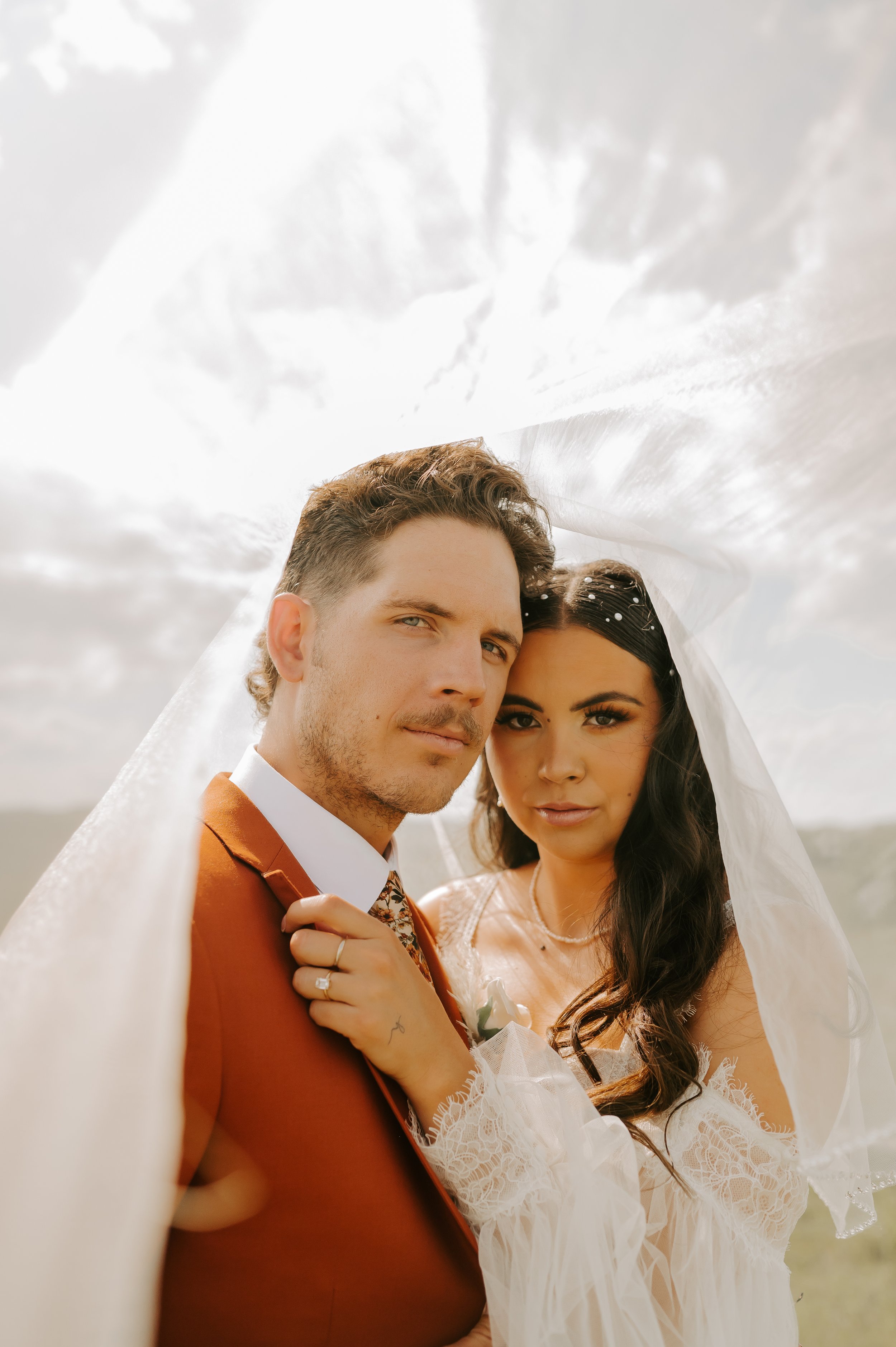 Bride and groom embrace under veil outdoors.