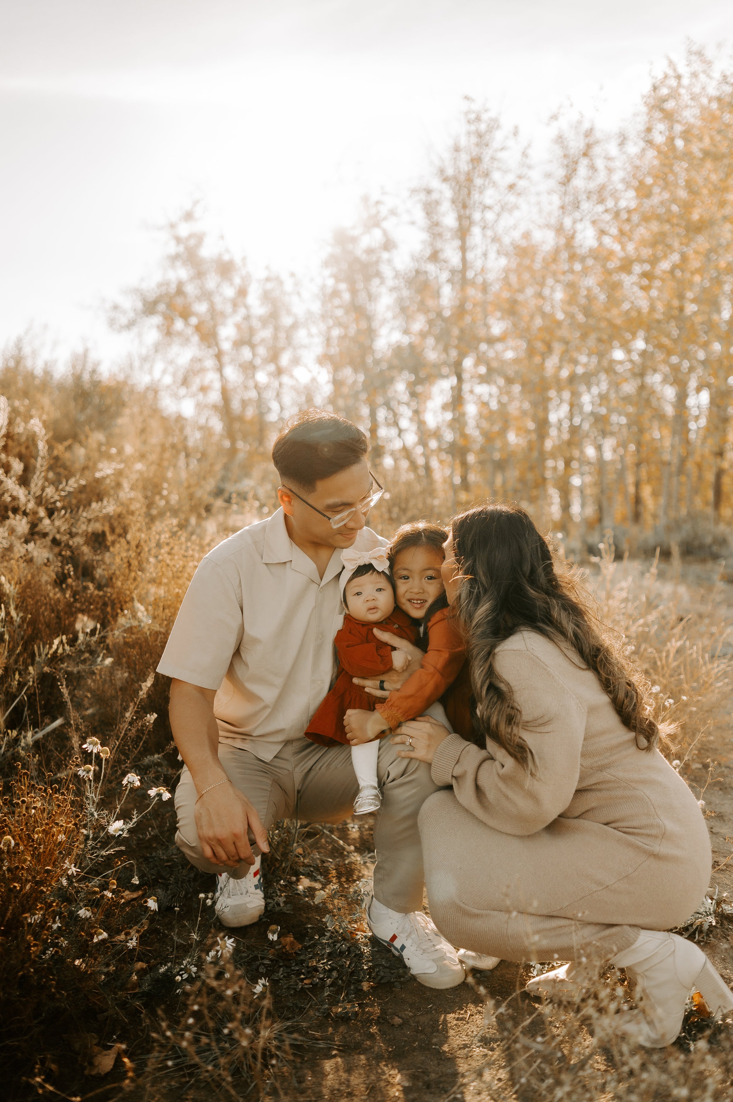 A family of four in a field during autumn, with trees in the background. The parents are crouching and smiling at their two young children, who are wearing red outfits. The scene is warm and softly lit.