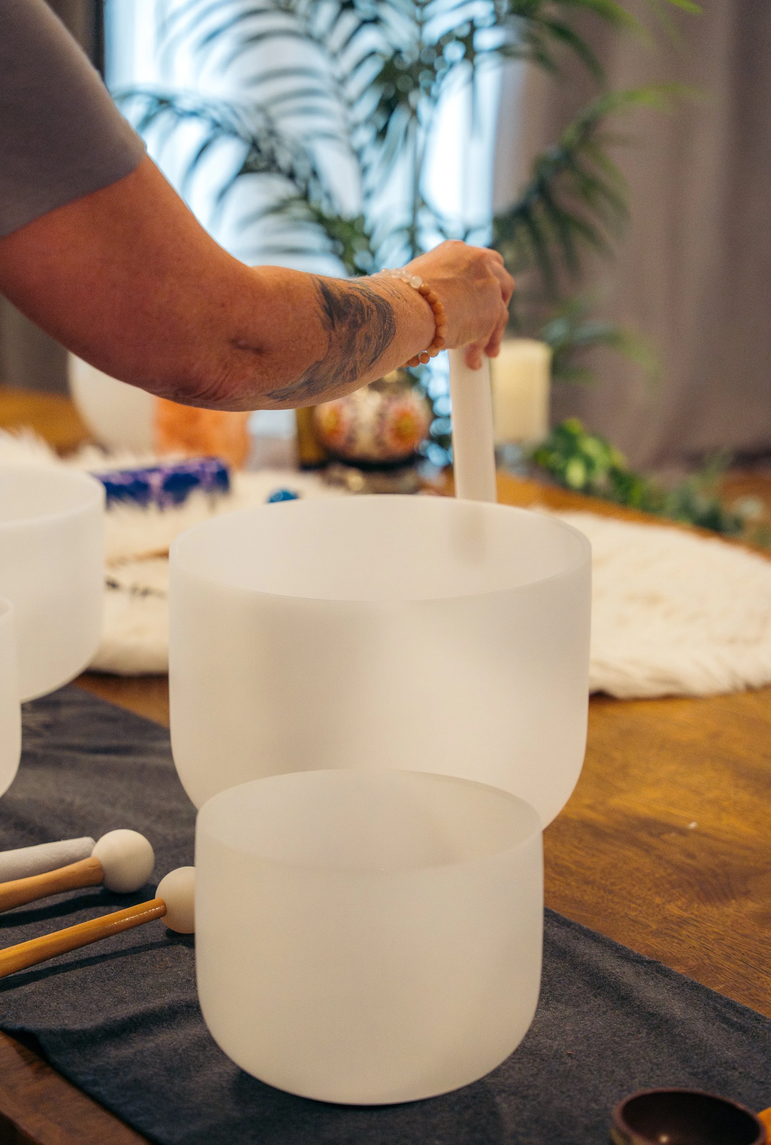Person playing a white crystal singing bowl with a mallet on a wooden table decorated with other musical instruments and plants in the background.