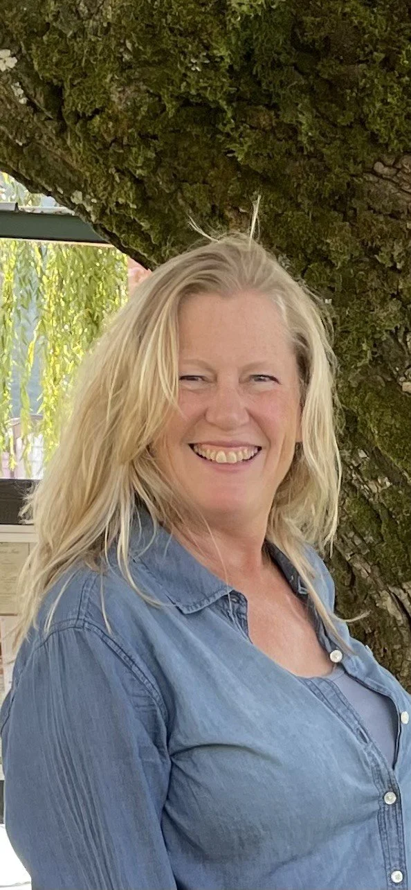 A smiling blonde woman standing outdoors next to a large tree with textured bark, in front of a window with greenery outside.