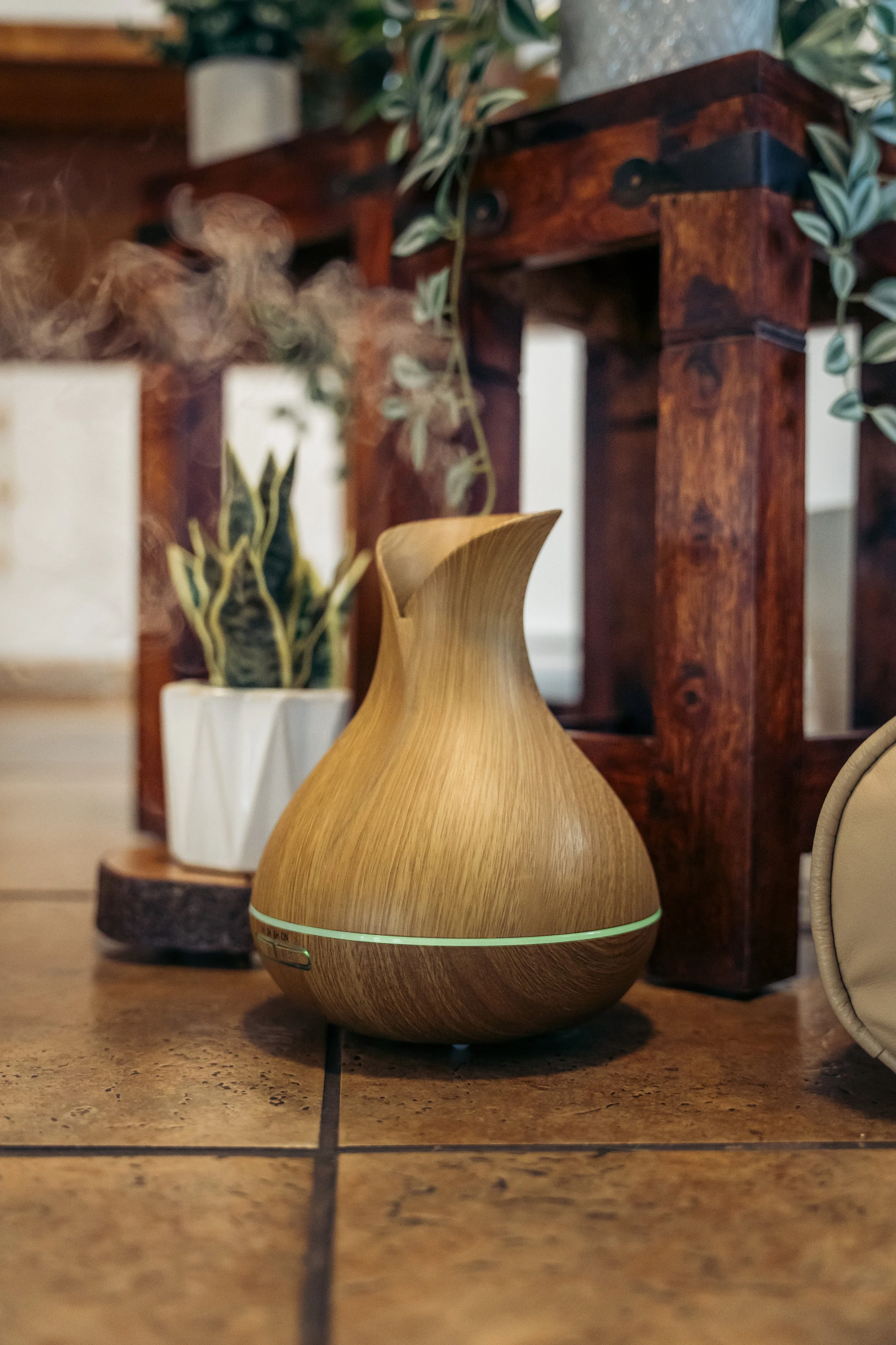 Wooden diffuser with smoke coming out, placed on a tiled floor in front of a wooden cabinet and potted plants.