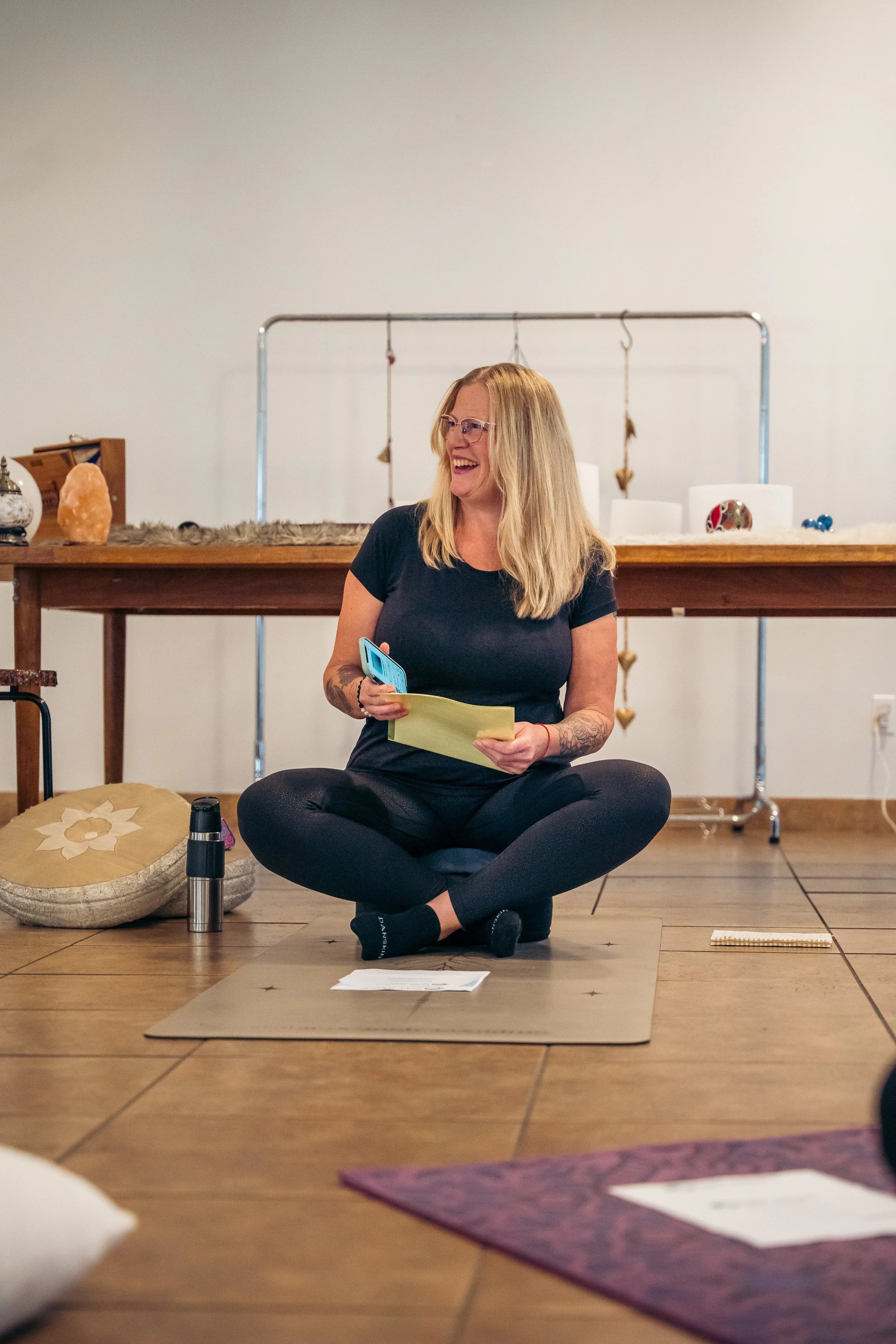 Woman sitting cross-legged on a yoga mat, smiling, holding a yellow paper and a phone in a room with wooden floor and minimal decor.
