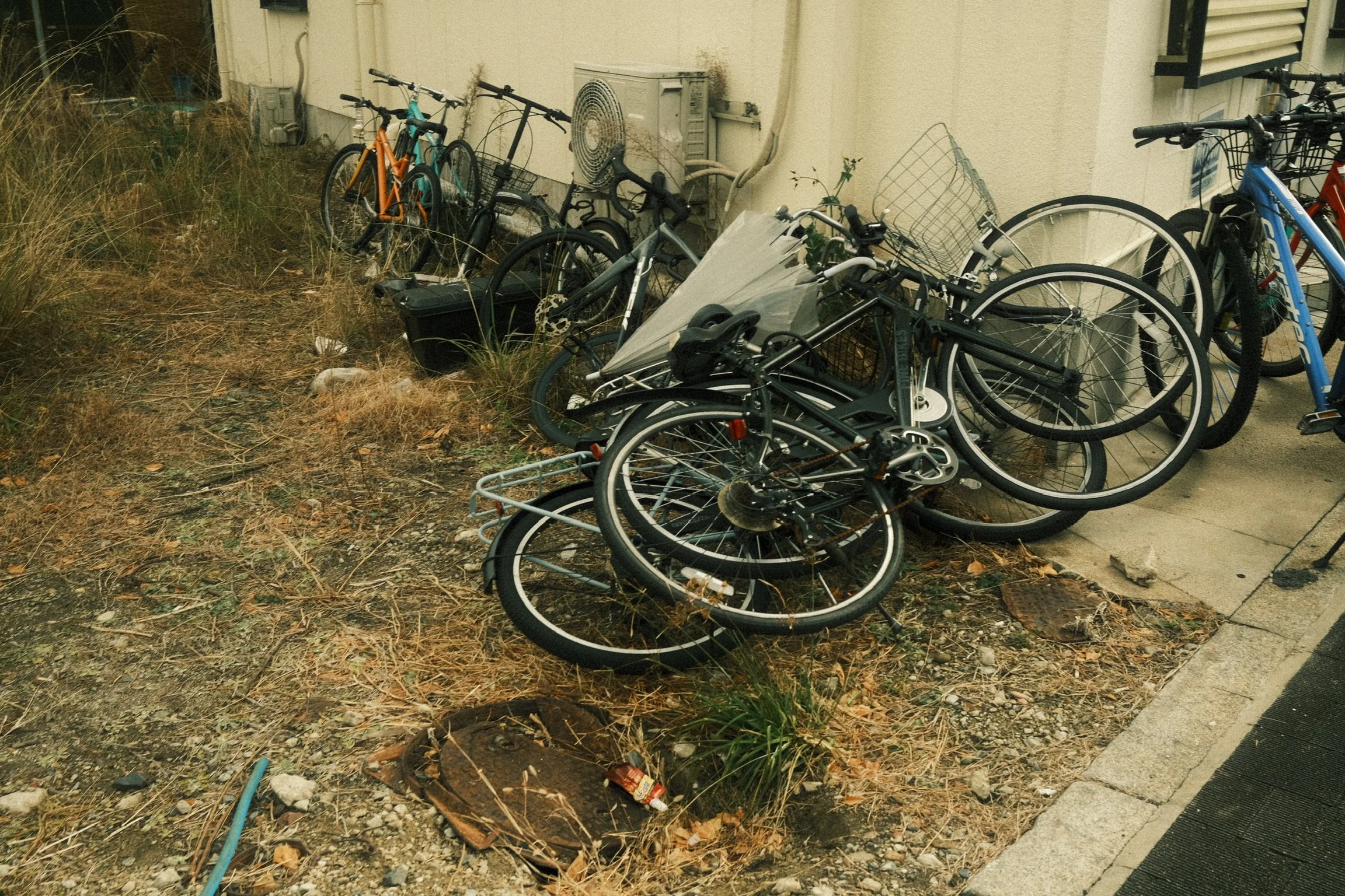 A cluttered outdoor area adjacent to a building with multiple bicycles, some parked upright and others on the ground, along with a small pile of trash, dry grass, and debris.