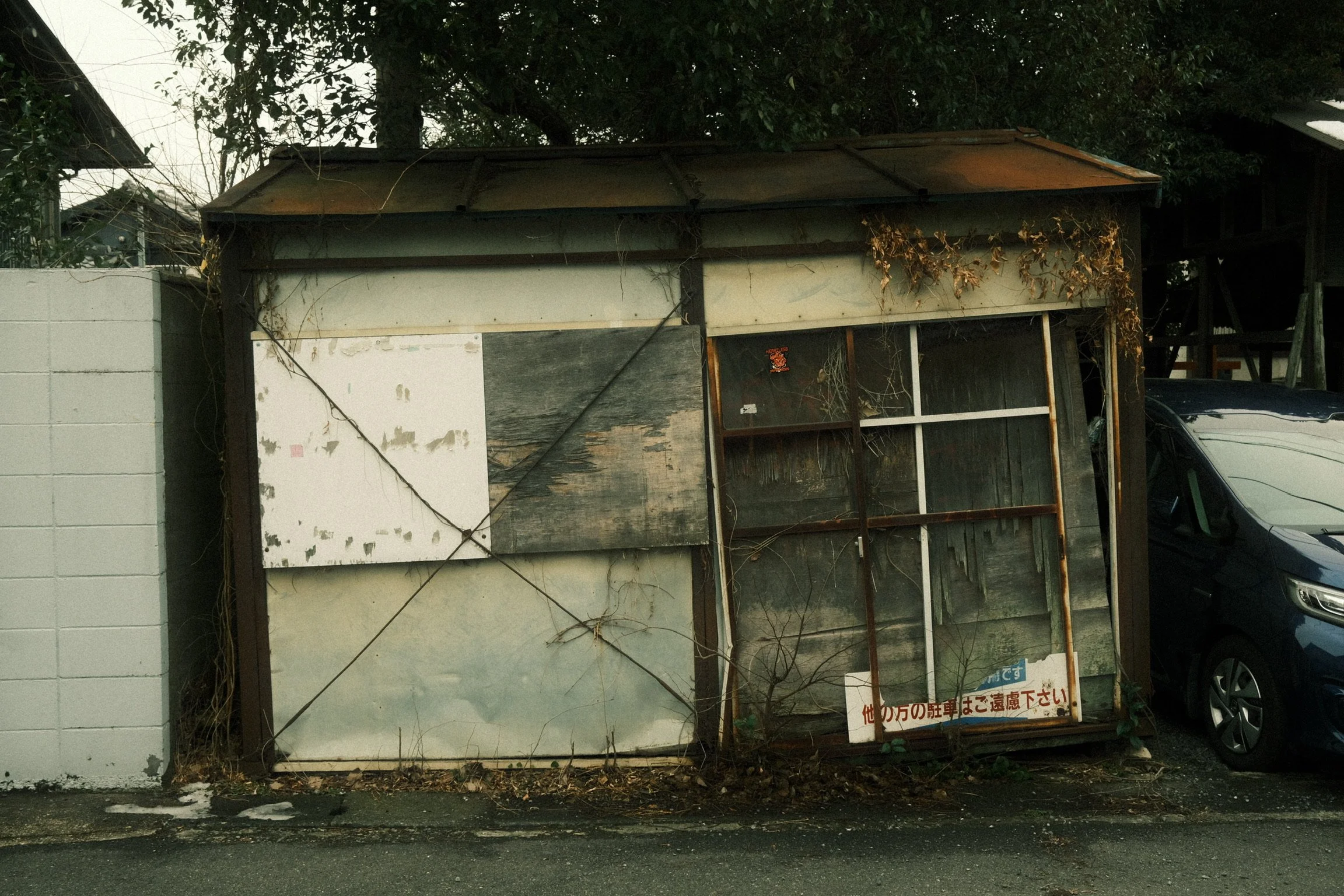An old, dilapidated wooden structure with rusty metal framing, overgrown with dried vines, situated next to a white concrete wall and partially obscured by a black car.