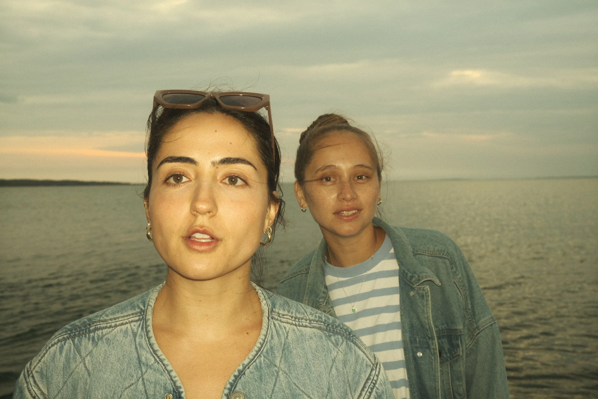 Two young women standing by a body of water during sunset, wearing denim jackets. One woman is wearing sunglasses on her head, and they are both looking toward the camera with relaxed expressions.
