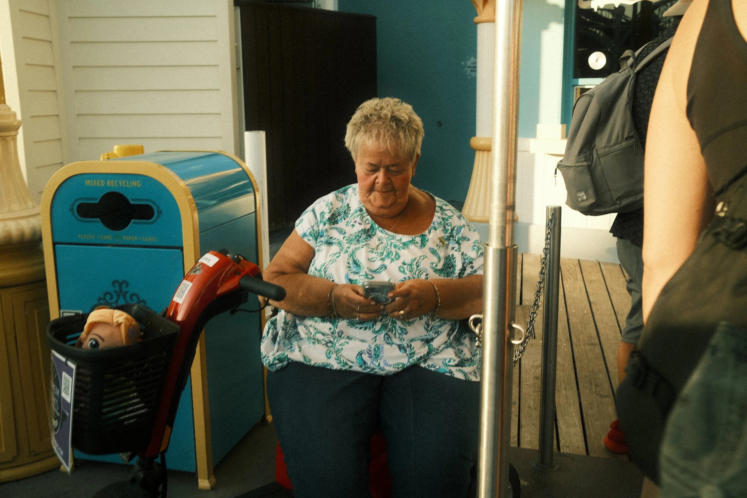 An elderly woman with short gray hair wearing a white and green patterned shirt, sitting and looking at her phone. She is inside a building near a recycling bin labeled mixed recycling, which contains various items including a bag with a toy doll's f