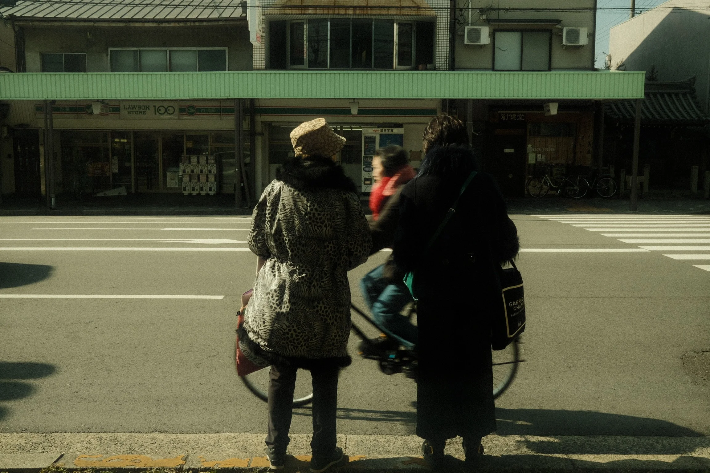 Three women standing on a city sidewalk in front of a store with a green awning. One woman is in a leopard-print coat and wide-brimmed hat, another wears a dark coat and has a bag, and the third woman, slightly blurred, has a red scarf. A bicycle is 