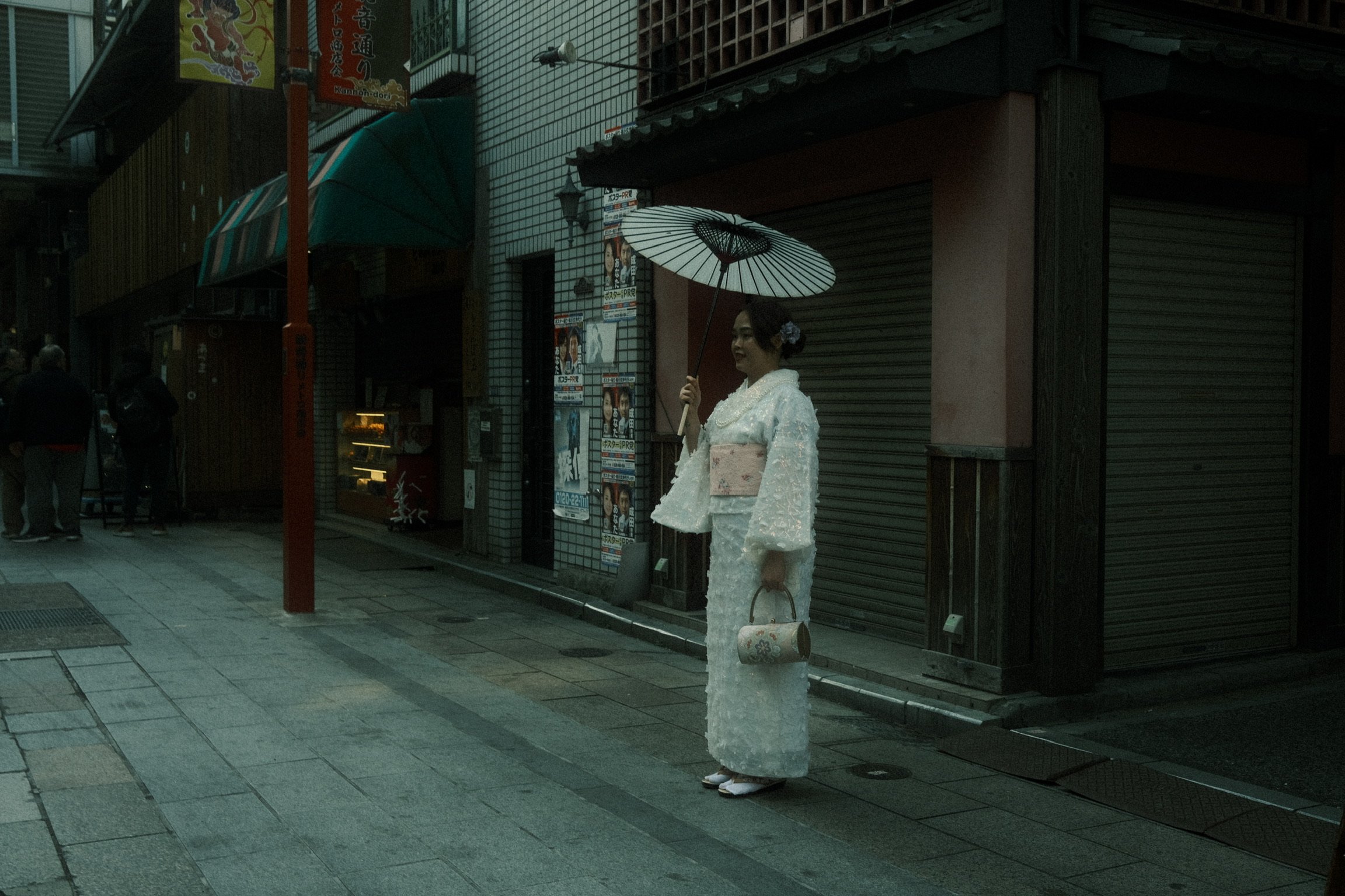 A woman wearing a traditional Japanese kimono holding a parasol and a handbag, standing on a city street at dusk.