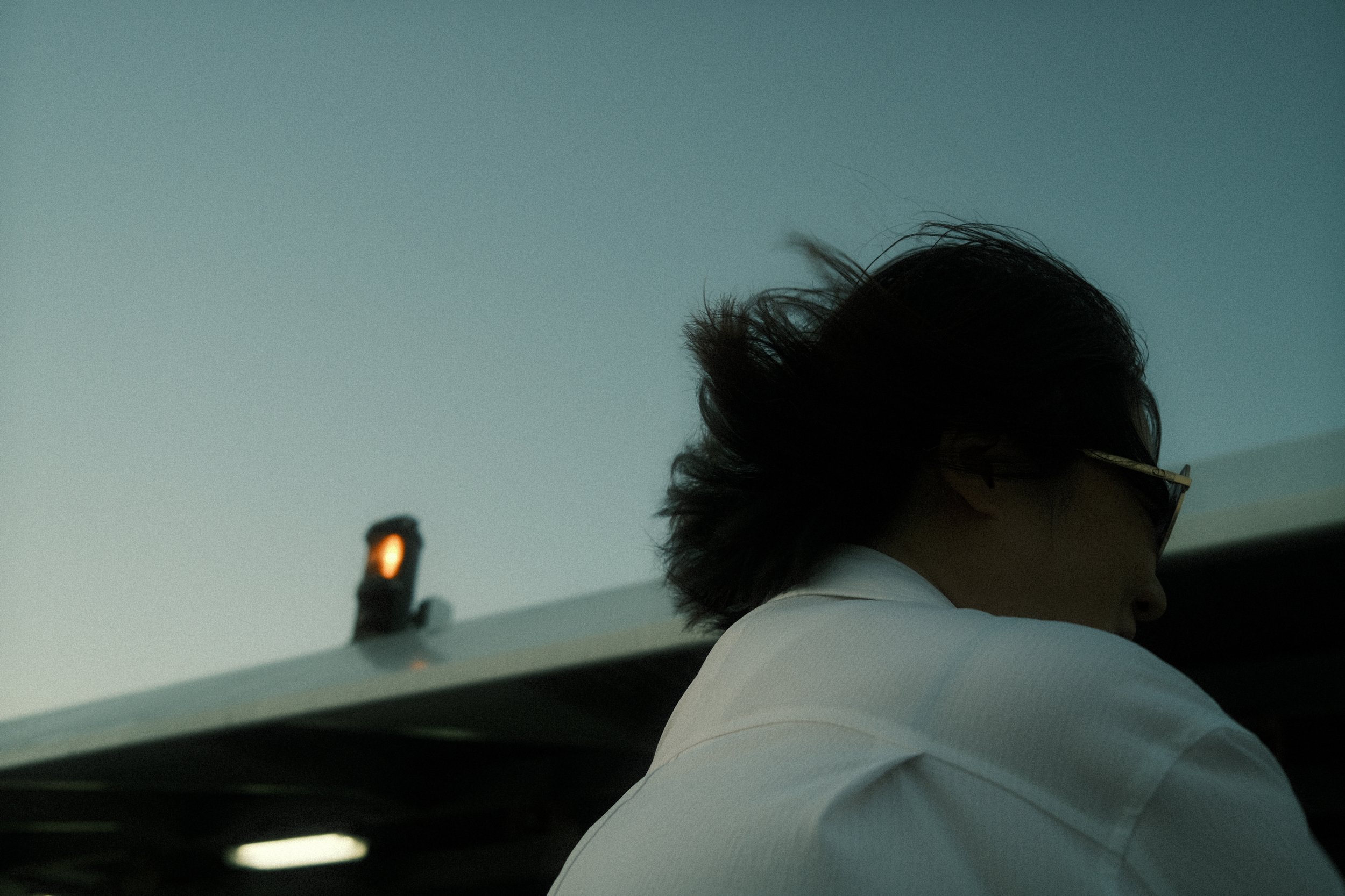 Partial view of a person with dark, tousled hair, wearing glasses, captured from behind, sitting inside an aircraft during twilight.