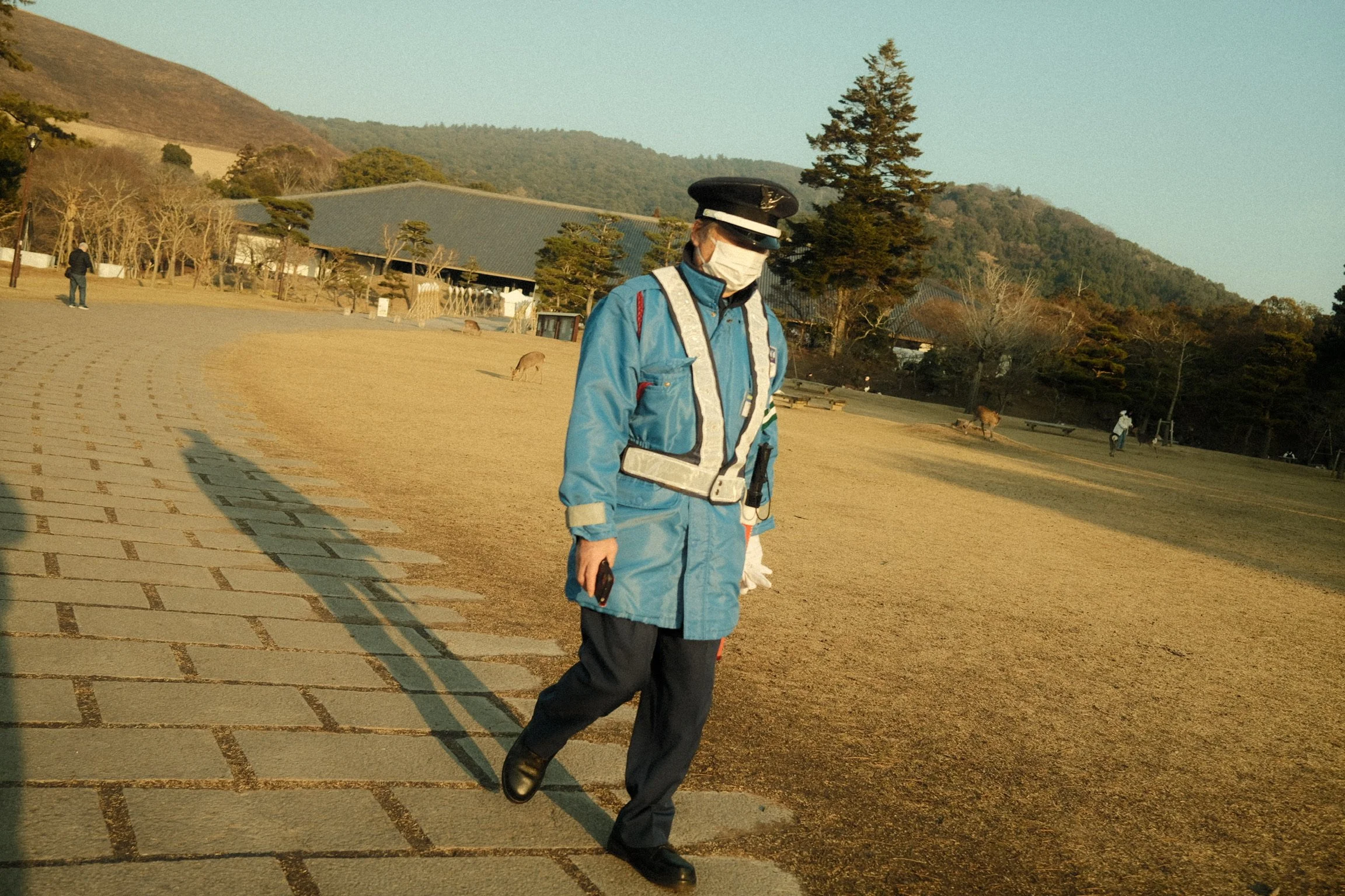 A person dressed as a police officer walking outdoors in a park with trees and hills in the background, wearing a face mask and sunglasses.