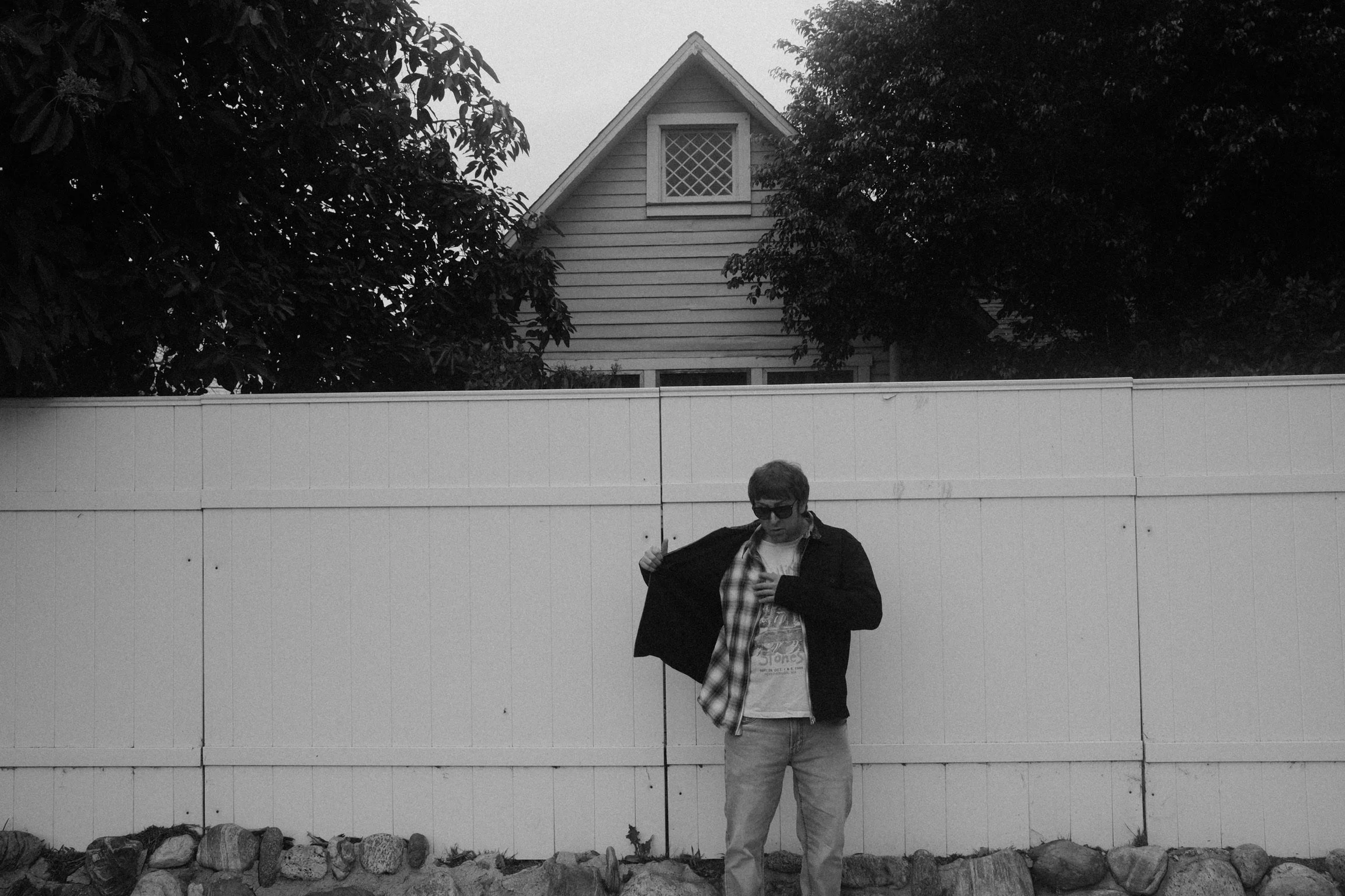 A young man with sunglasses adjusting his jacket while standing in front of a white fence and a garden bed with rocks. Behind him is a house with a gabled roof and two windows, surrounded by trees.