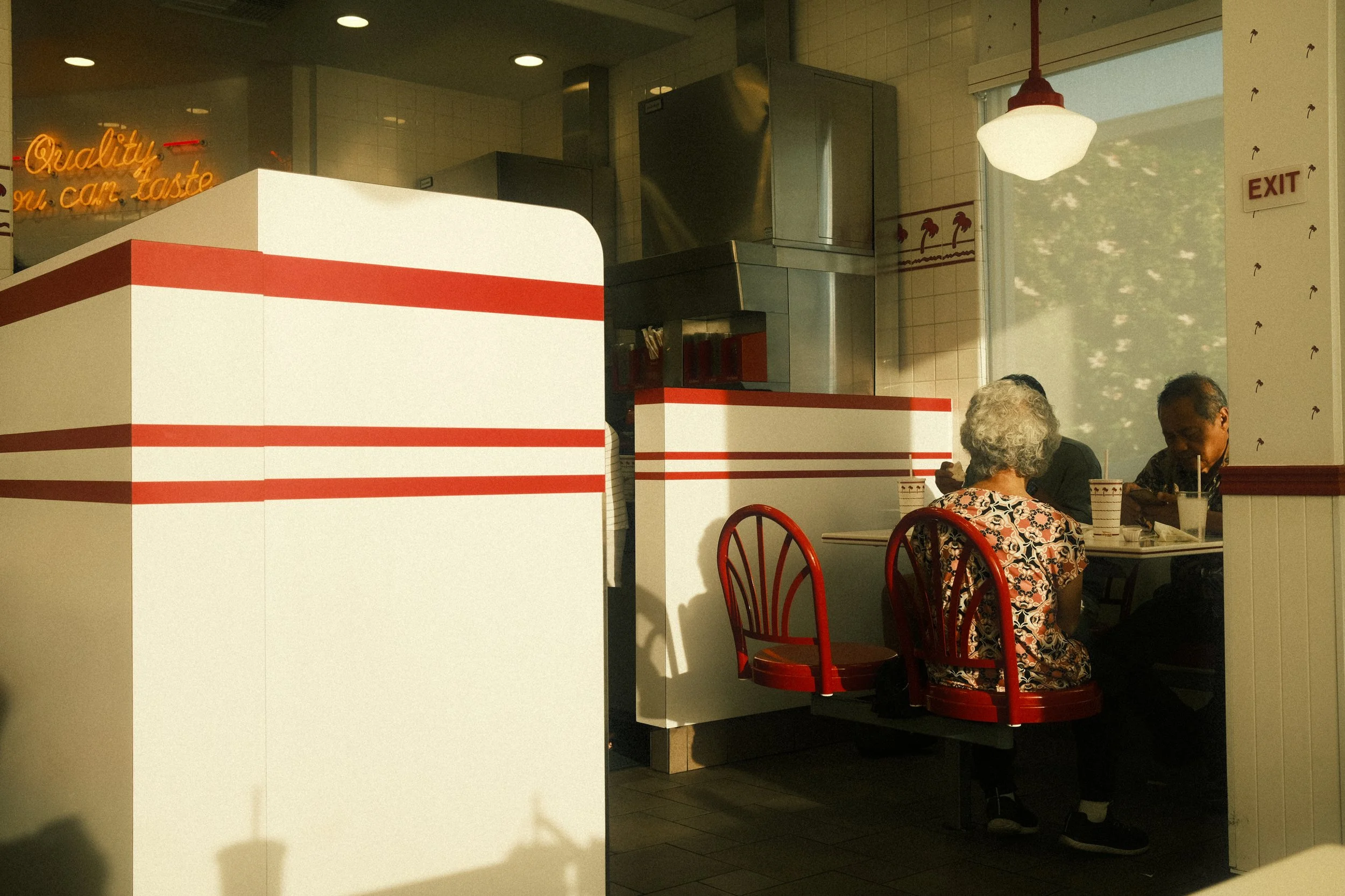Inside a fast-food restaurant with white and red decor, two elderly people are sitting at a table with red chairs, eating and drinking. A large window with a view of trees is in the background, and a sign on the wall reads "exit."