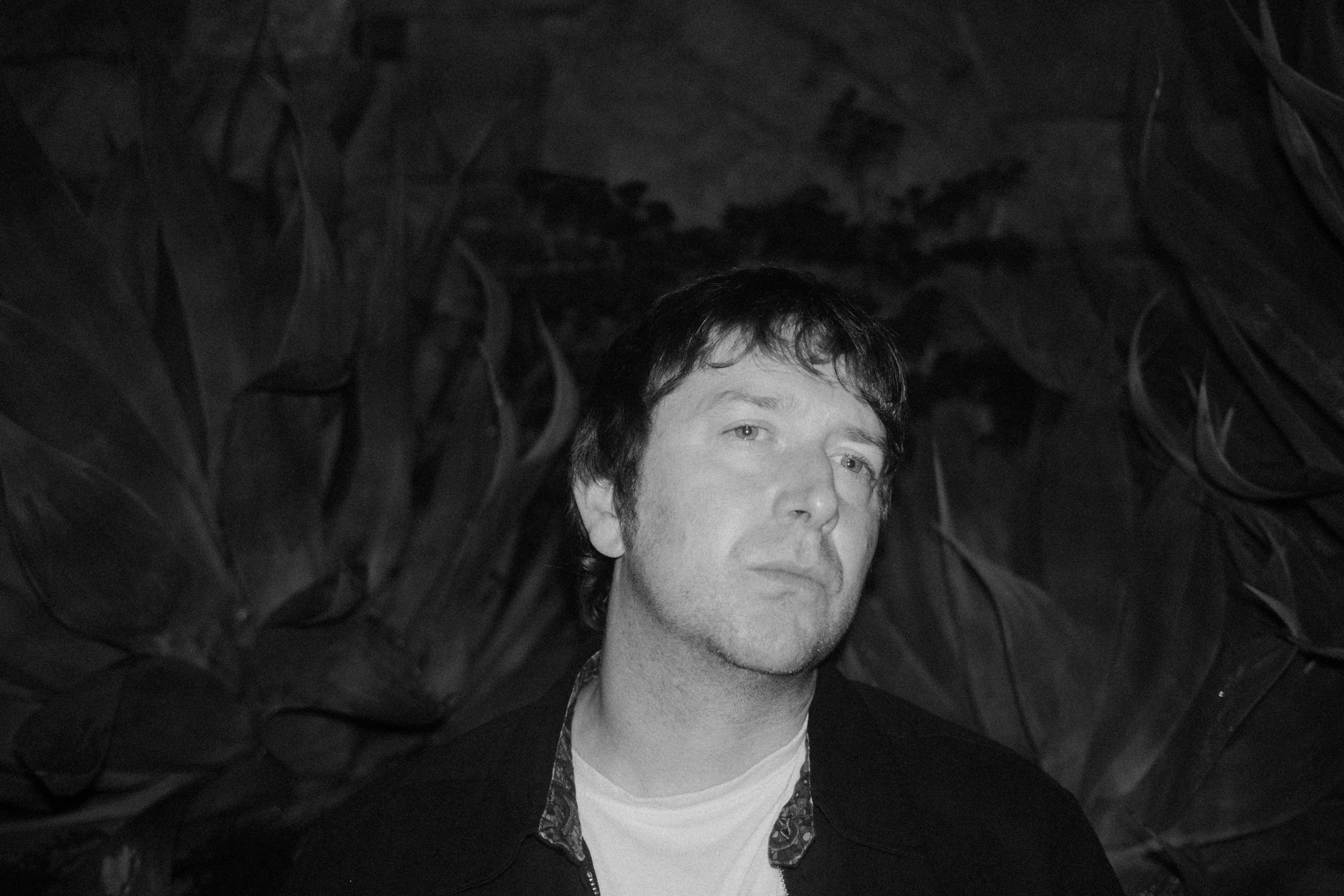 Black and white photo of a man with dark hair and light-colored eyes, looking slightly away from the camera, with a serious expression. He is wearing a dark jacket over a light shirt, with an ornate pattern on the inside collar, and is surrounded by 