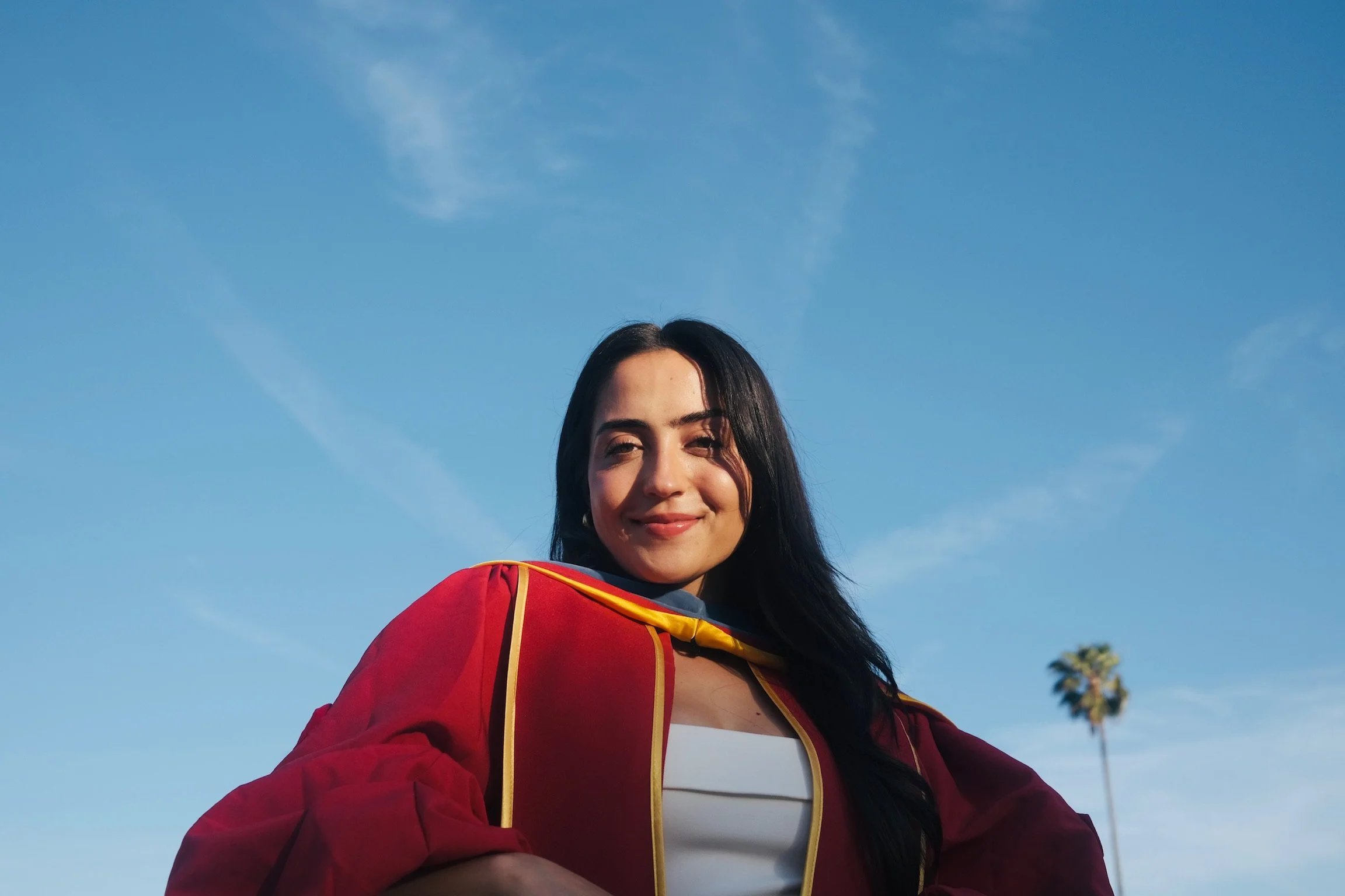 A young woman wearing a red graduation gown and mortarboard smiling outdoors against a blue sky with a few clouds and a palm tree in the background.