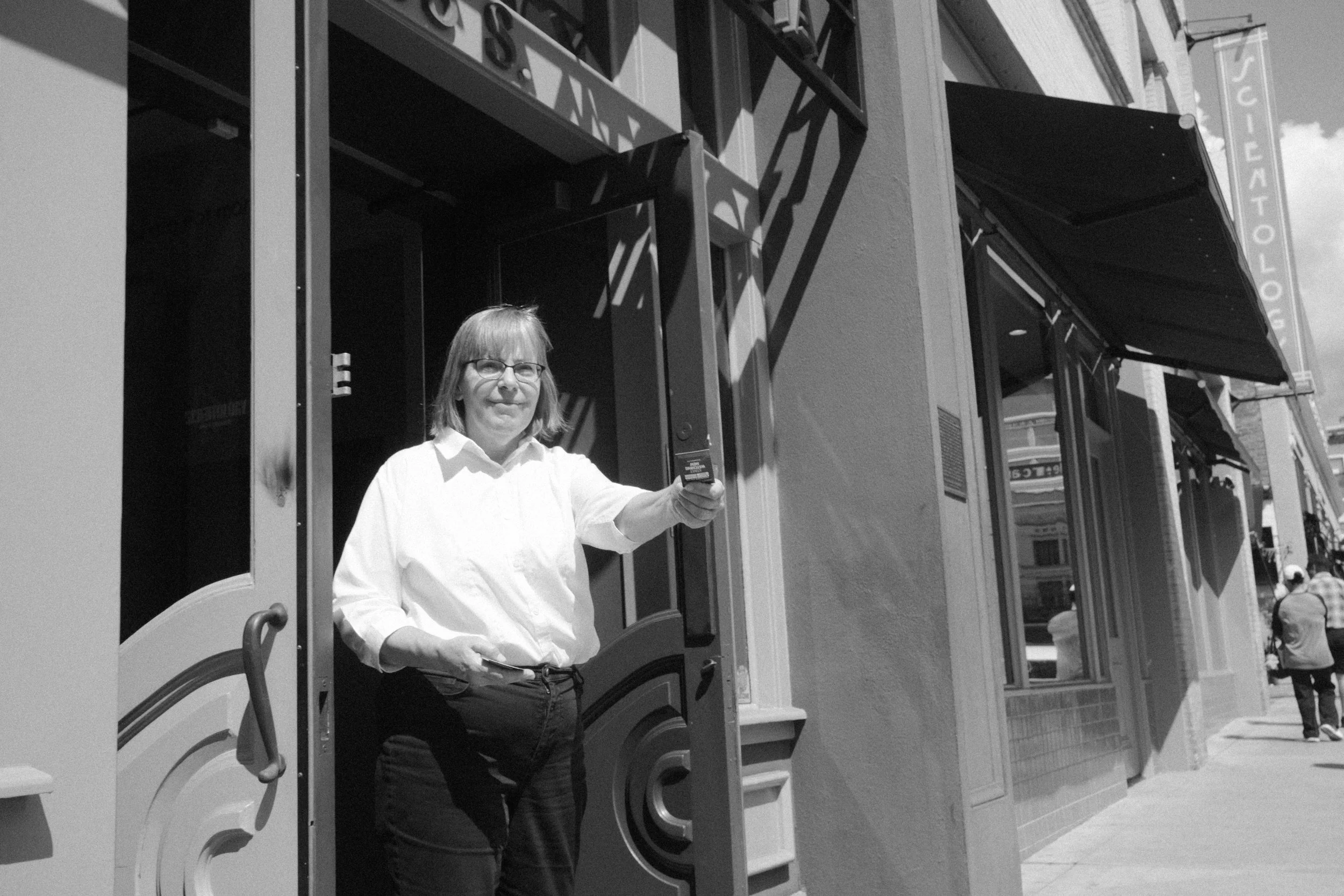 A woman with glasses and a light-colored shirt standing outside a storefront, holding a card or object in her hand. The storefront has awnings and window displays.