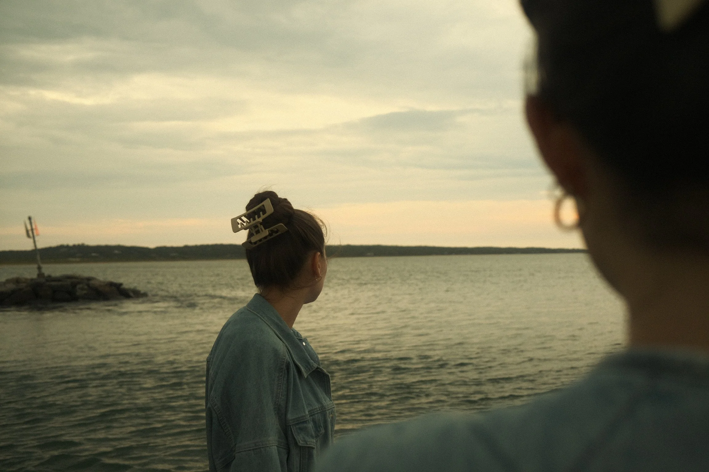 A woman with a large hair clip standing on a waterfront during sunset, with a person partly out of focus in the foreground, and a lighthouse in the distance.