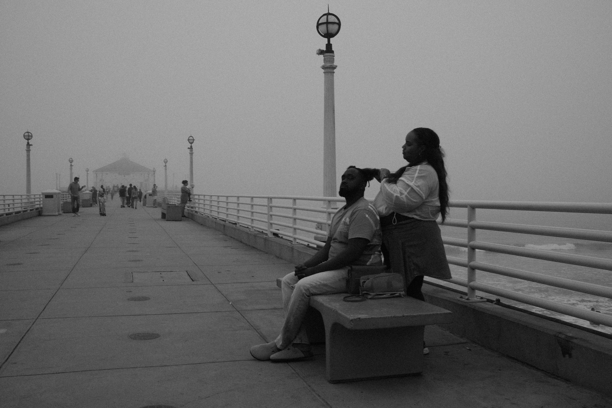 A woman styling a man's dreadlocks on a seaside pier with lamp posts and a covered pavilion in the background, during dusk or dawn.