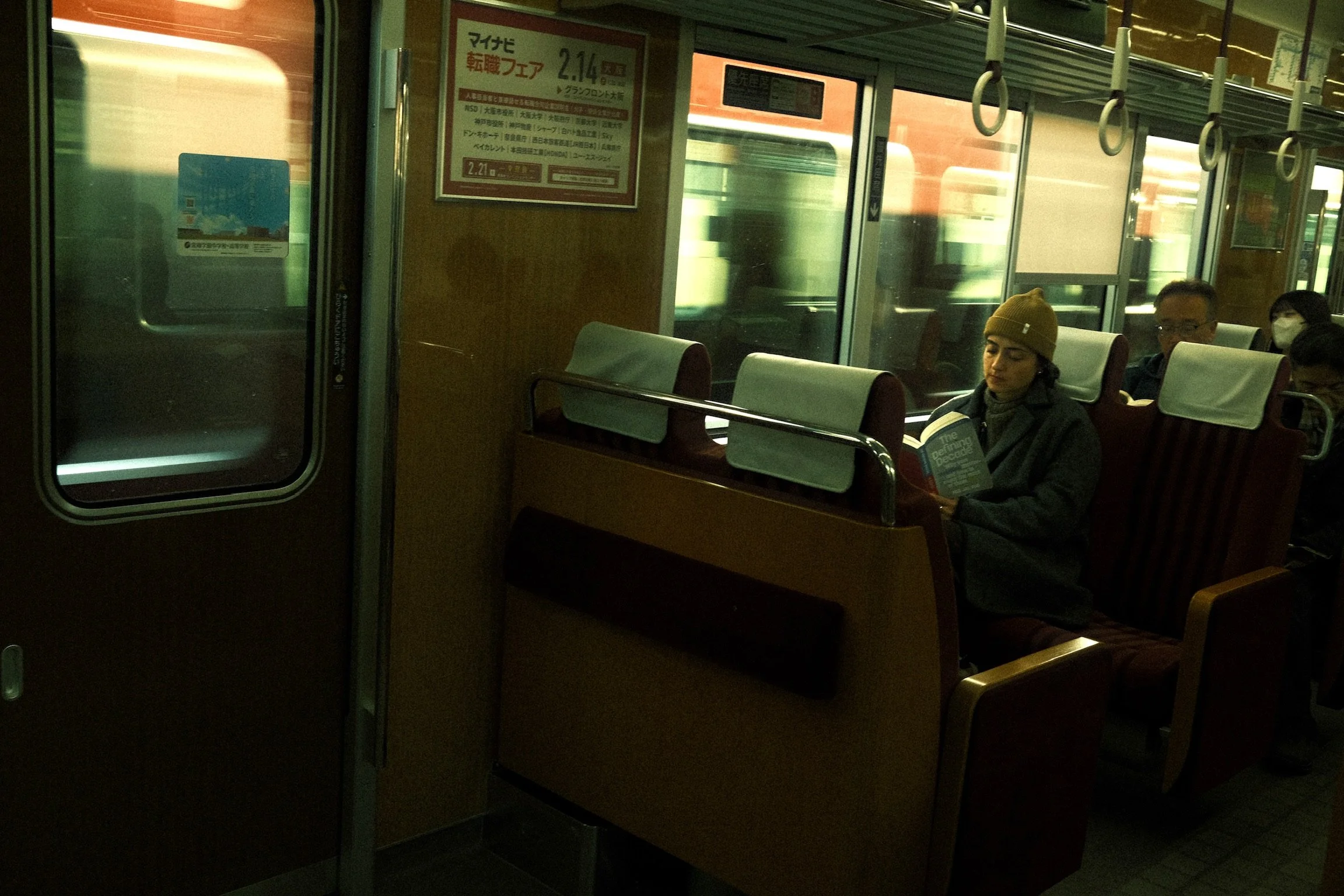 Inside a train with passengers seated, including a woman reading a book, and a window showing motion blur of outside scenery. There is also a sign with Japanese text.