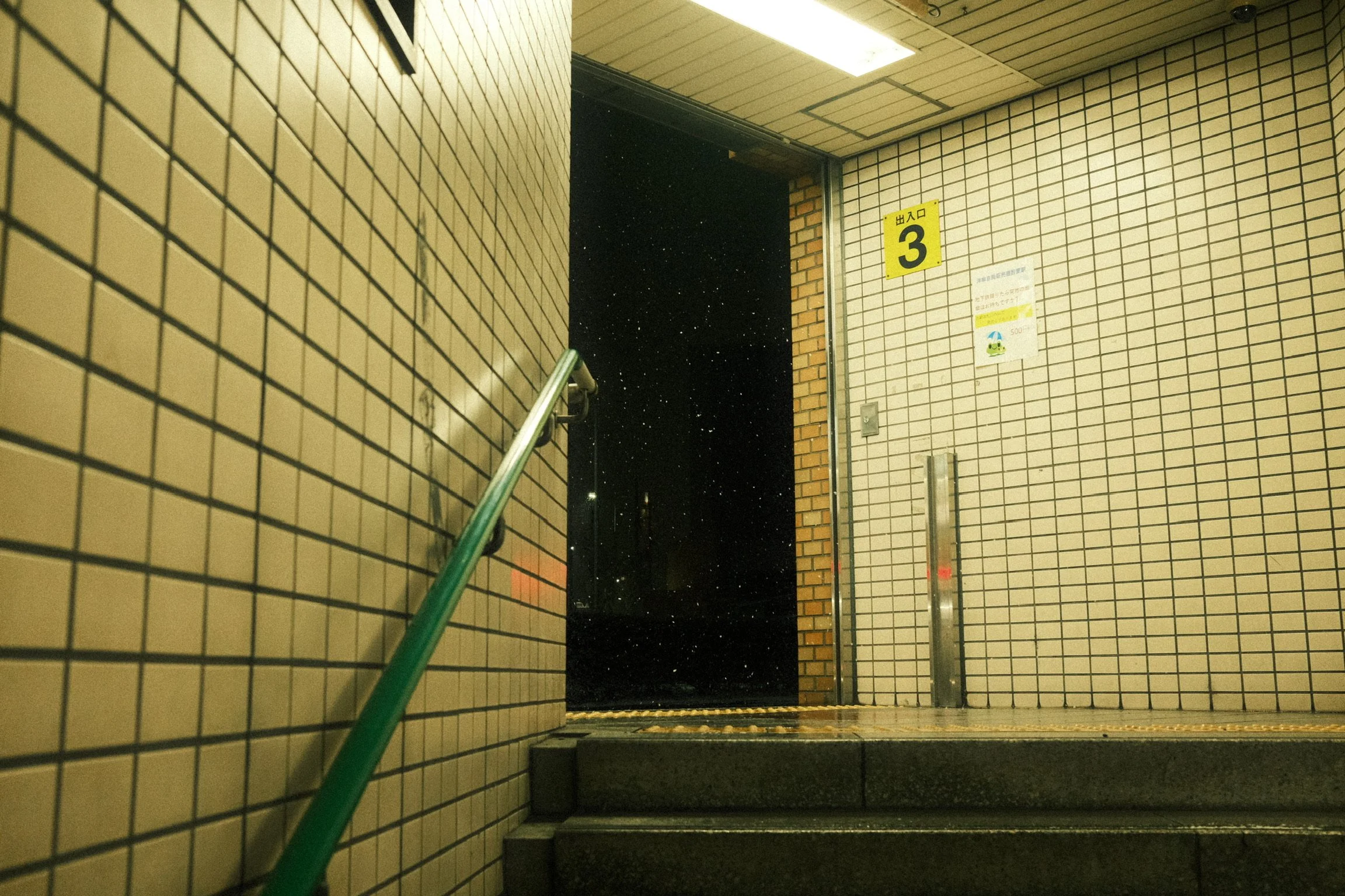 An indoor staircase entrance with beige tiled walls, a green handrail on the left side, a yellow sign with the number 3 on the wall, and a door opening to a dark nighttime sky visible outside, with stars