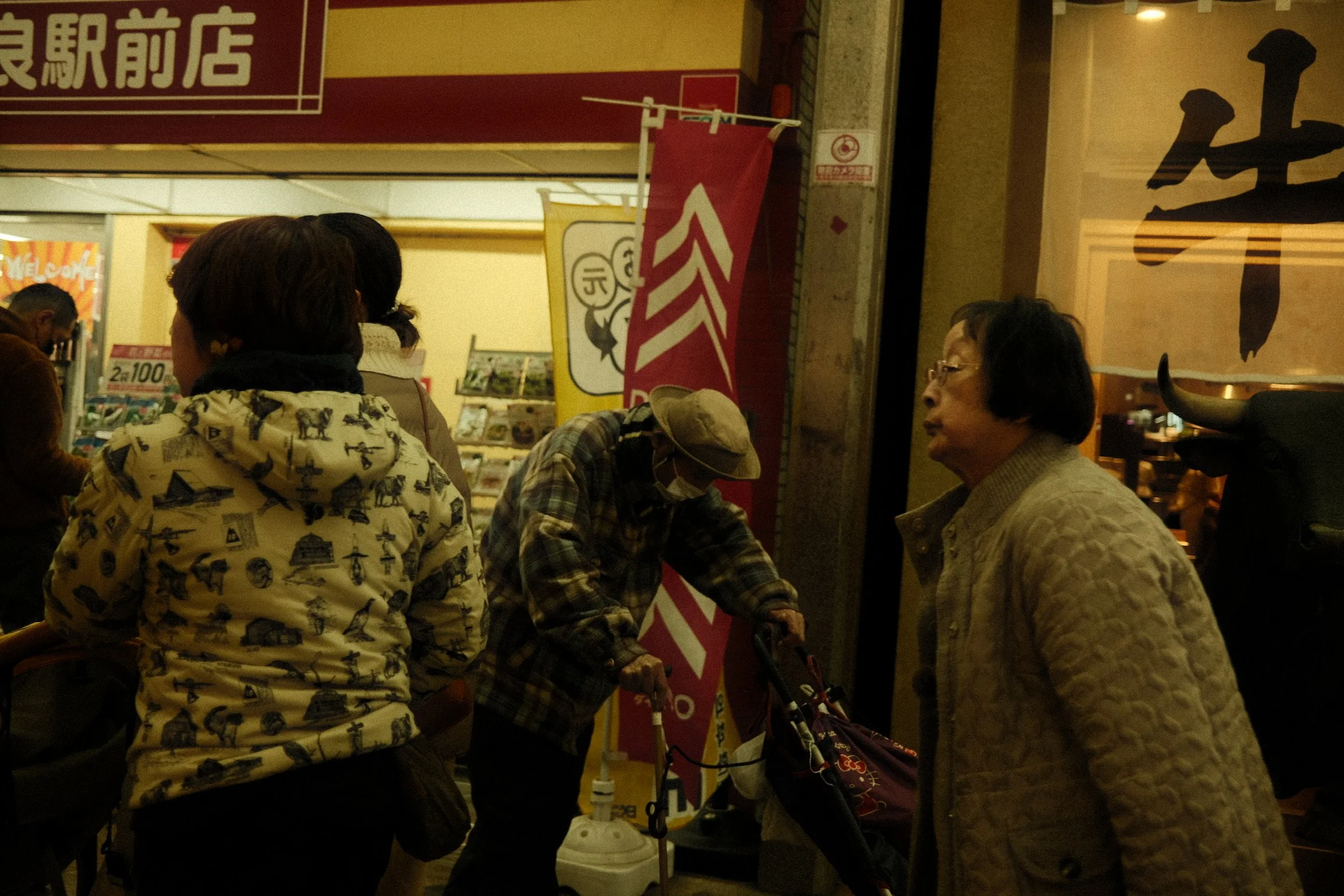 People standing and conversing inside a store or restaurant with Japanese signage, including a woman wearing a cream quilted jacket, an elderly woman with dark hair and glasses, and a person with a hat and a puffy jacket.