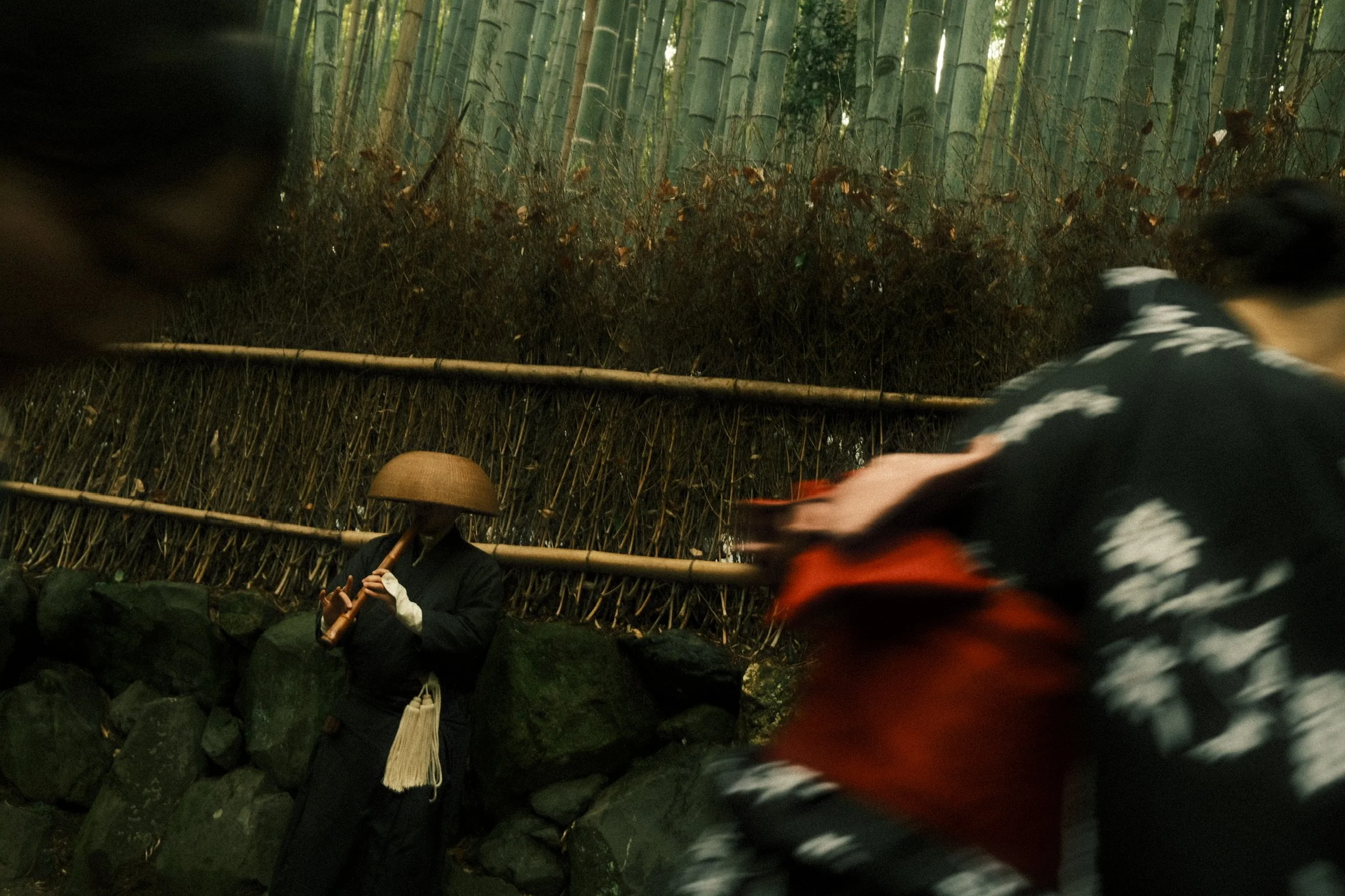 Two people in traditional clothing and bamboo hats are in a bamboo forest; one person is playing a flute, and the other is partially visible in motion.