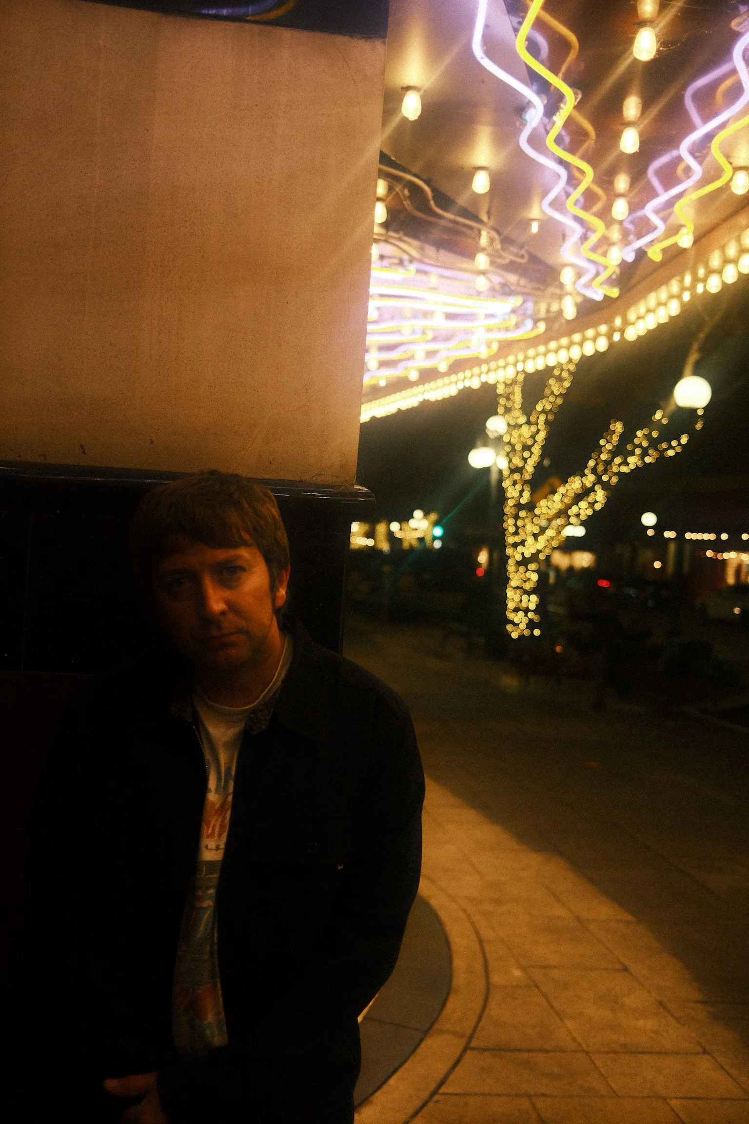 Night scene with decorative string lights, a young man with brown hair and a dark jacket standing near a column, illuminated by colorful neon lights on the ceiling, blurry city lights in the background.