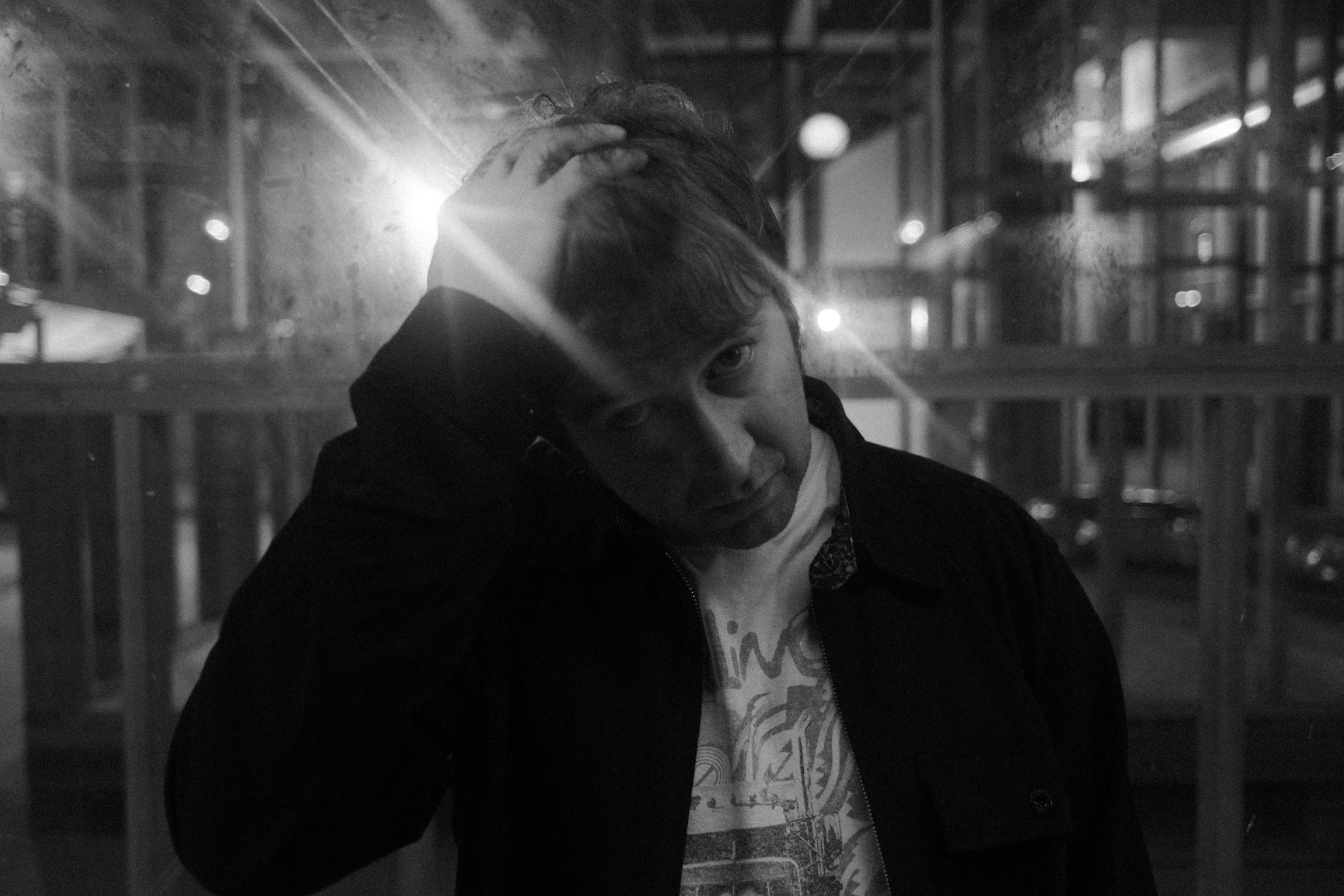 A young man with short hair, wearing a dark jacket and a t-shirt with a graphic design, stands outside at night and touches his forehead with one hand, appearing contemplative.
