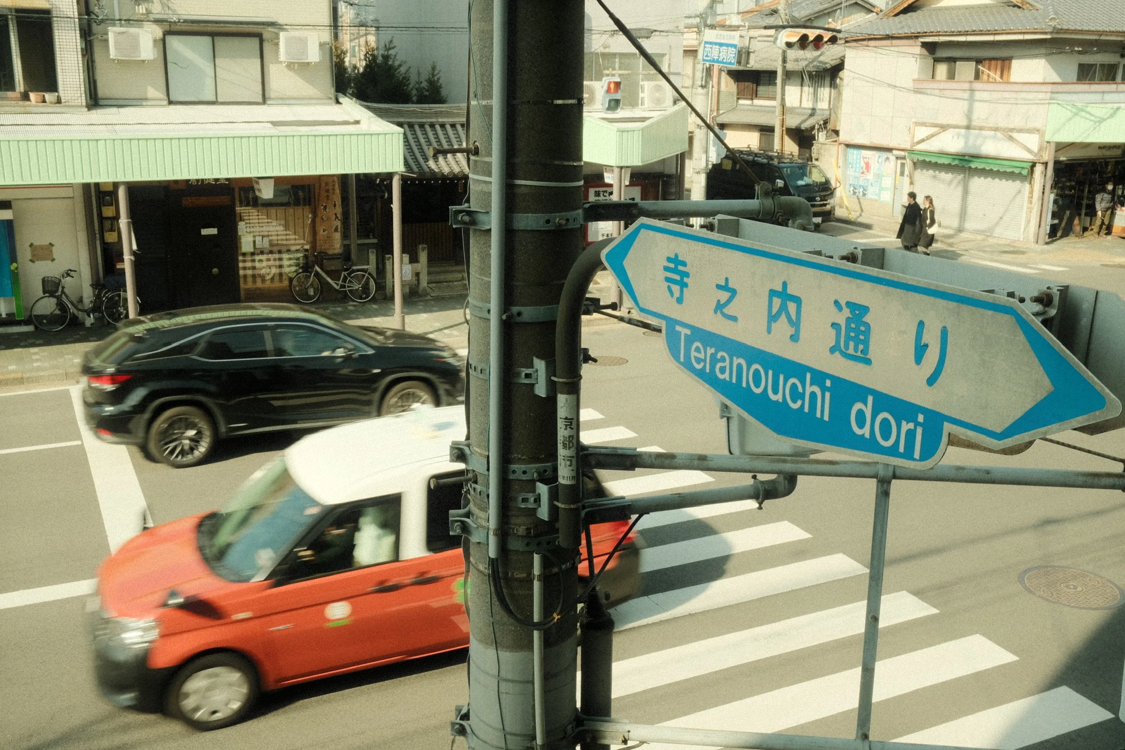 Street scene with cars, bicycles, pedestrians, and street signs in Japanese and English, taken from a window.