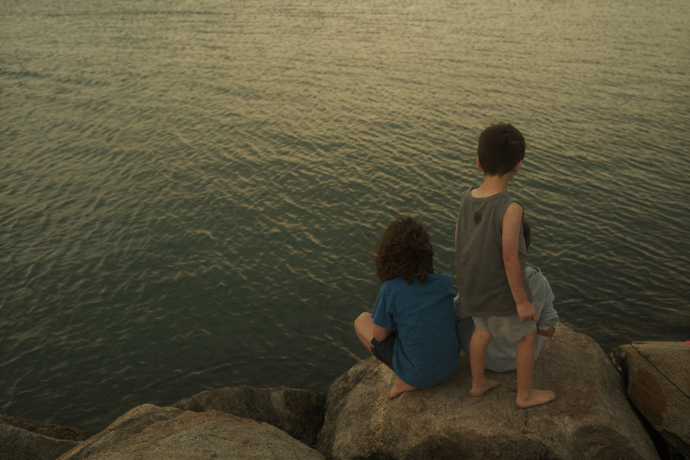 Three children sitting on rocks by a body of water, facing away from the camera, during sunset or dusk.