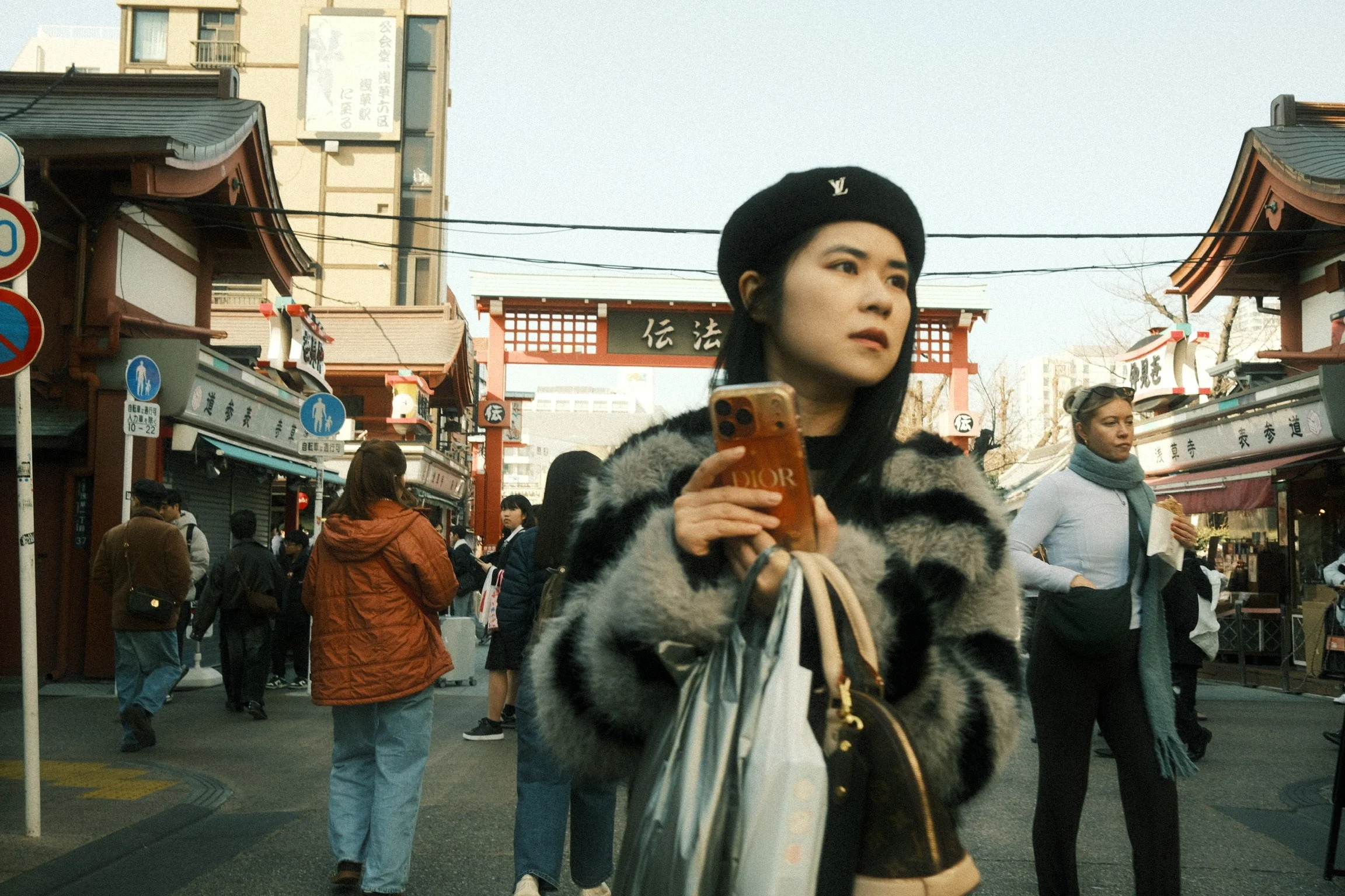 A woman in a black fur coat and black hat with a Louis Vuitton logo, holding a phone, standing in a busy street with people walking around, traditional Japanese temple gates in the background.