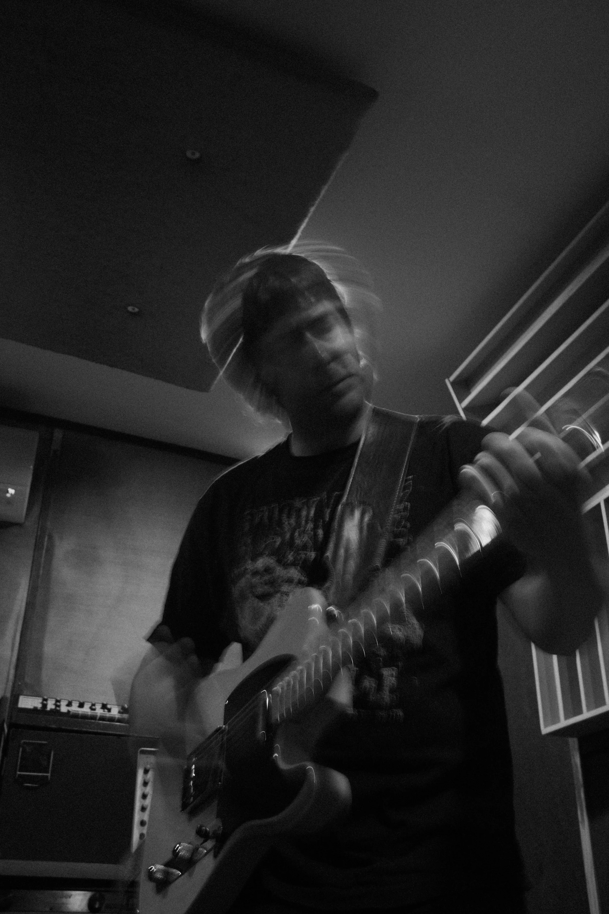 A person with light-colored, slightly messy hair is playing an electric guitar in a dimly lit room. The person is wearing a dark T-shirt and appears to be focusing on their playing. Music equipment, including a keyboard and audio equipment, can be se