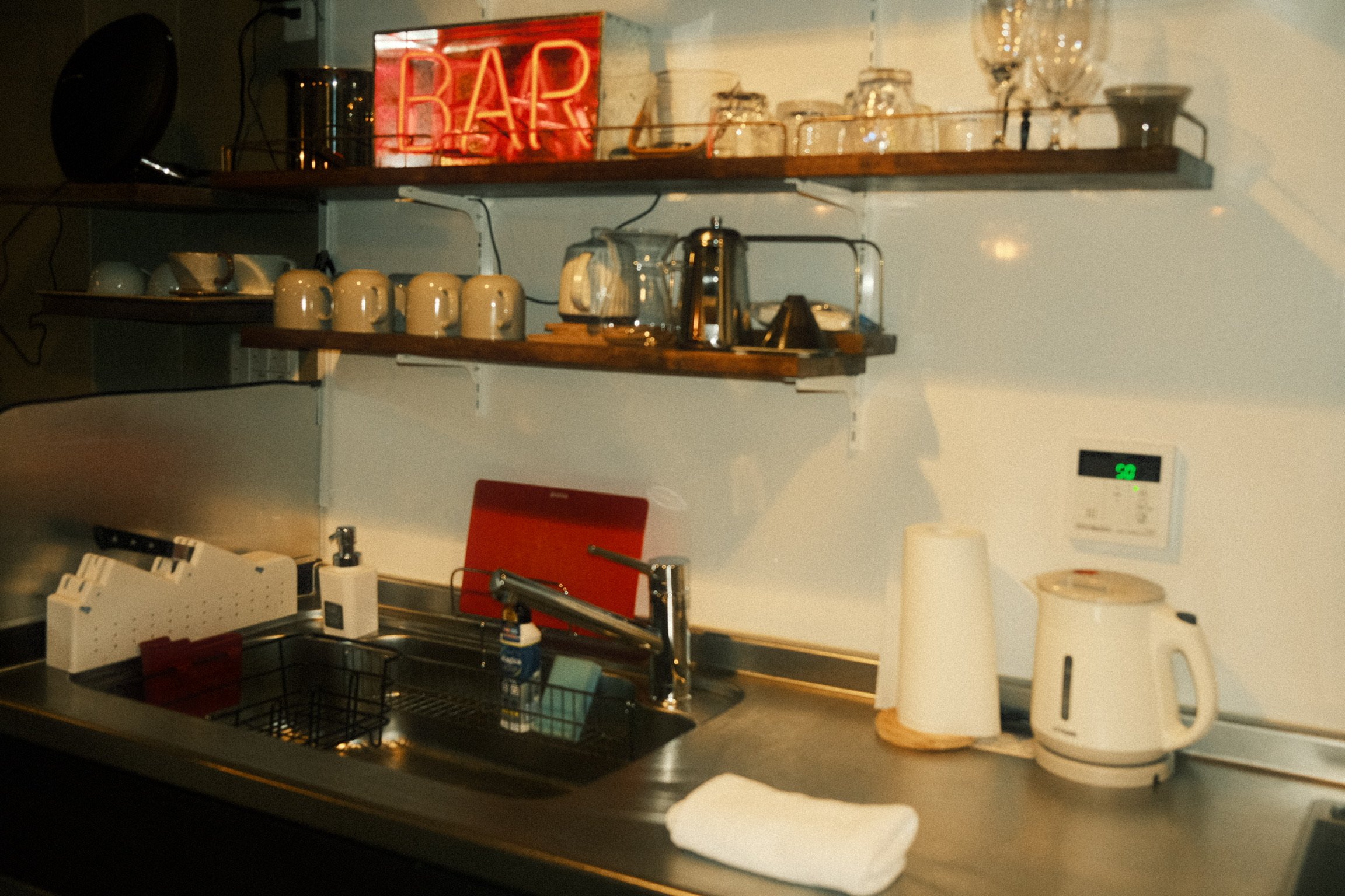 Kitchen countertop with various appliances including a microwave, electric kettle, paper towel dispenser, and pots. Wooden shelves hold cups, glassware, and a neon 'BAR' sign.
