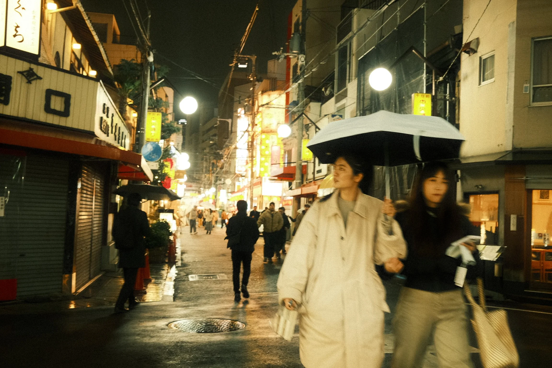 People walking with umbrellas on a rainy night in a busy city street illuminated by streetlights and signs.