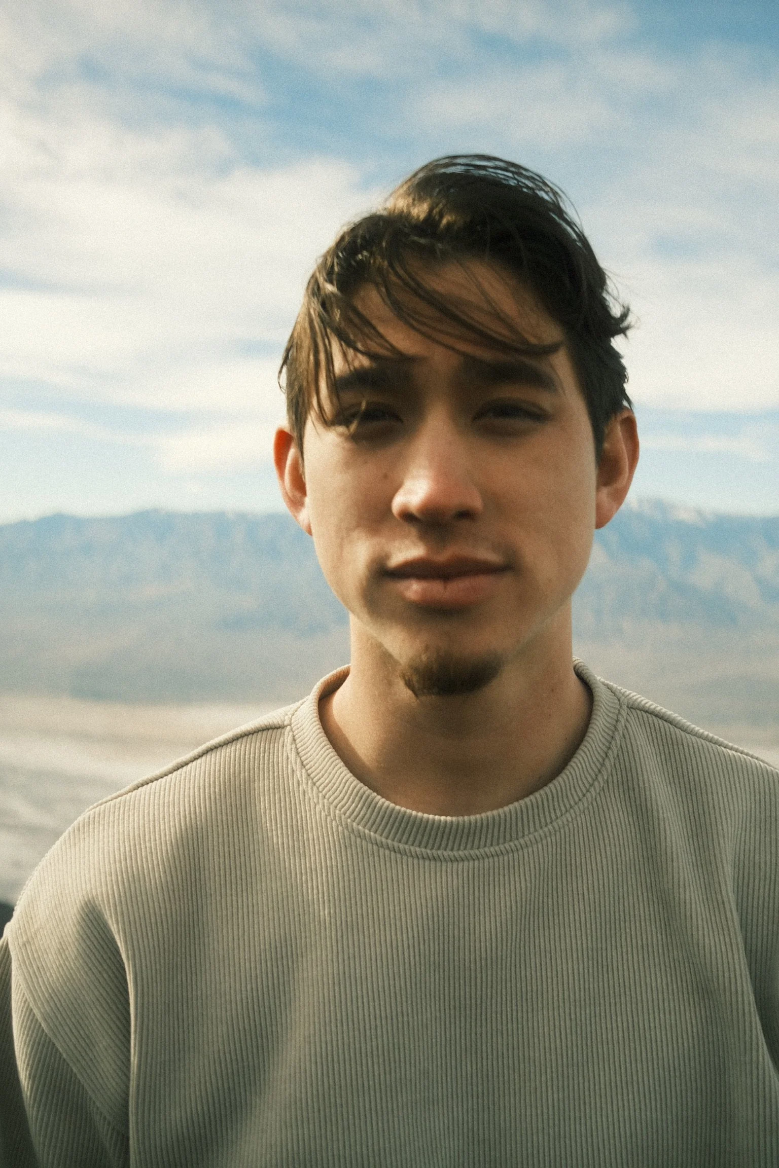 A young man with dark hair and a mustache wearing a beige ribbed long-sleeve shirt, standing outdoors against a mountain landscape and a partly cloudy sky.