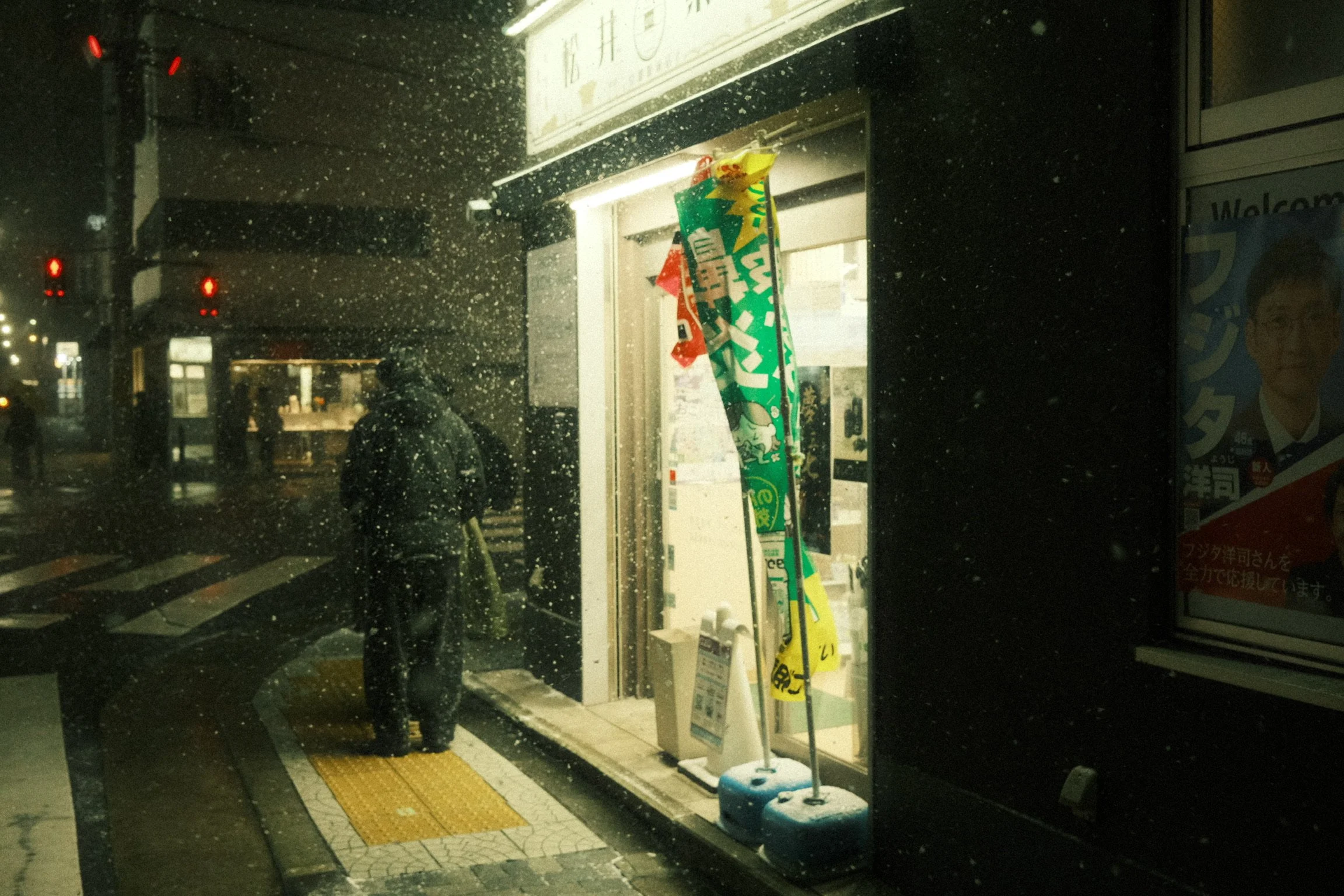 A rainy night scene outside a convenience store with a person standing at the entrance, illuminated by store lights, with advertisements and signs in Japanese, and specks of rain visible on a glass window.