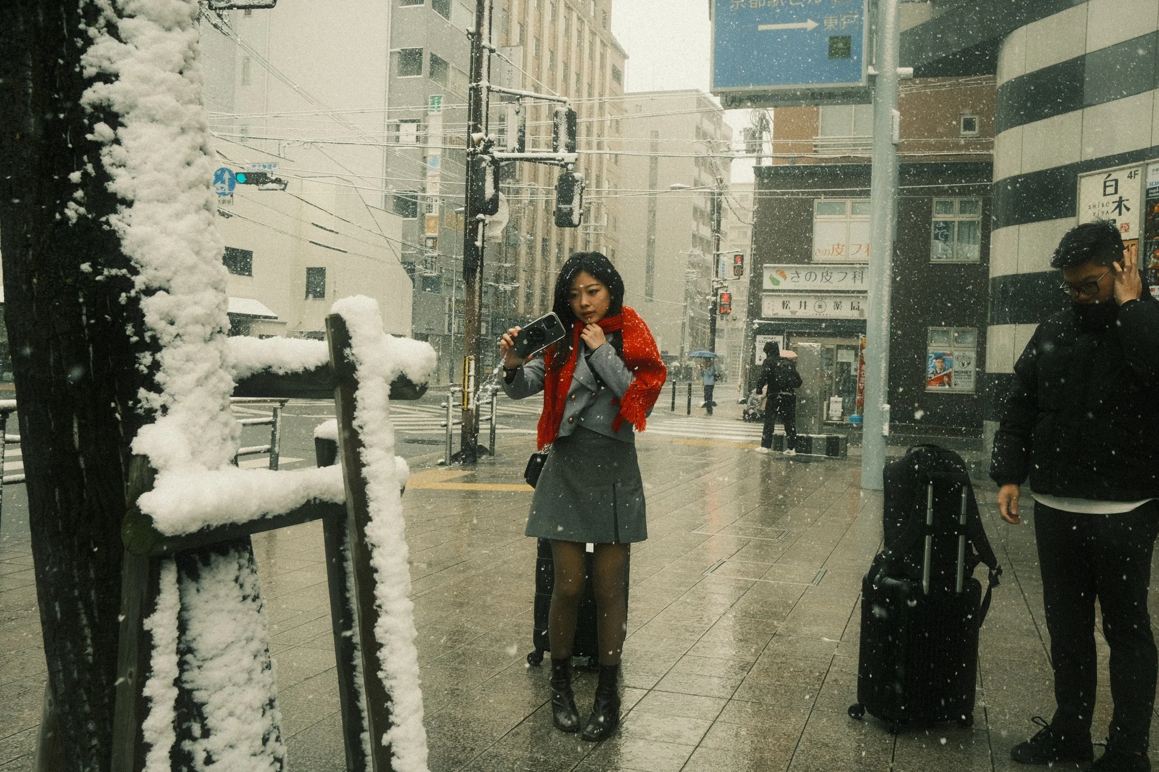 A woman in black tights, a gray skirt, and a red scarf is standing at a snowy city street corner, holding a purse and a camera. To her right, a man in glasses and a black coat is adjusting his ear. Snow covers a bench and trees, with city buildings, 