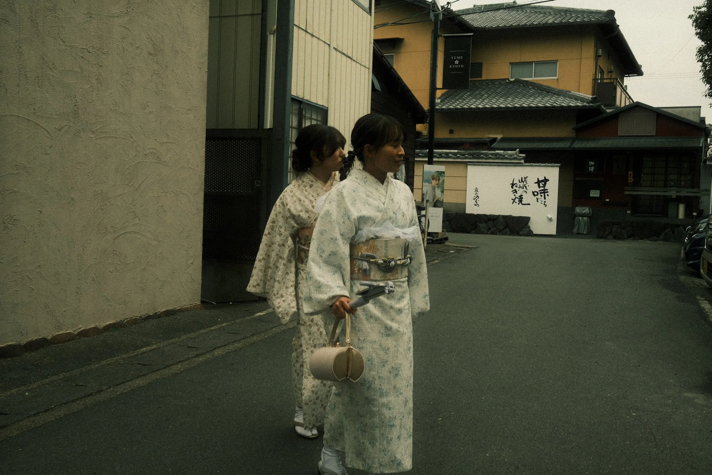 Two women dressed in traditional Japanese kimonos walking on a street, with buildings and signage in the background.