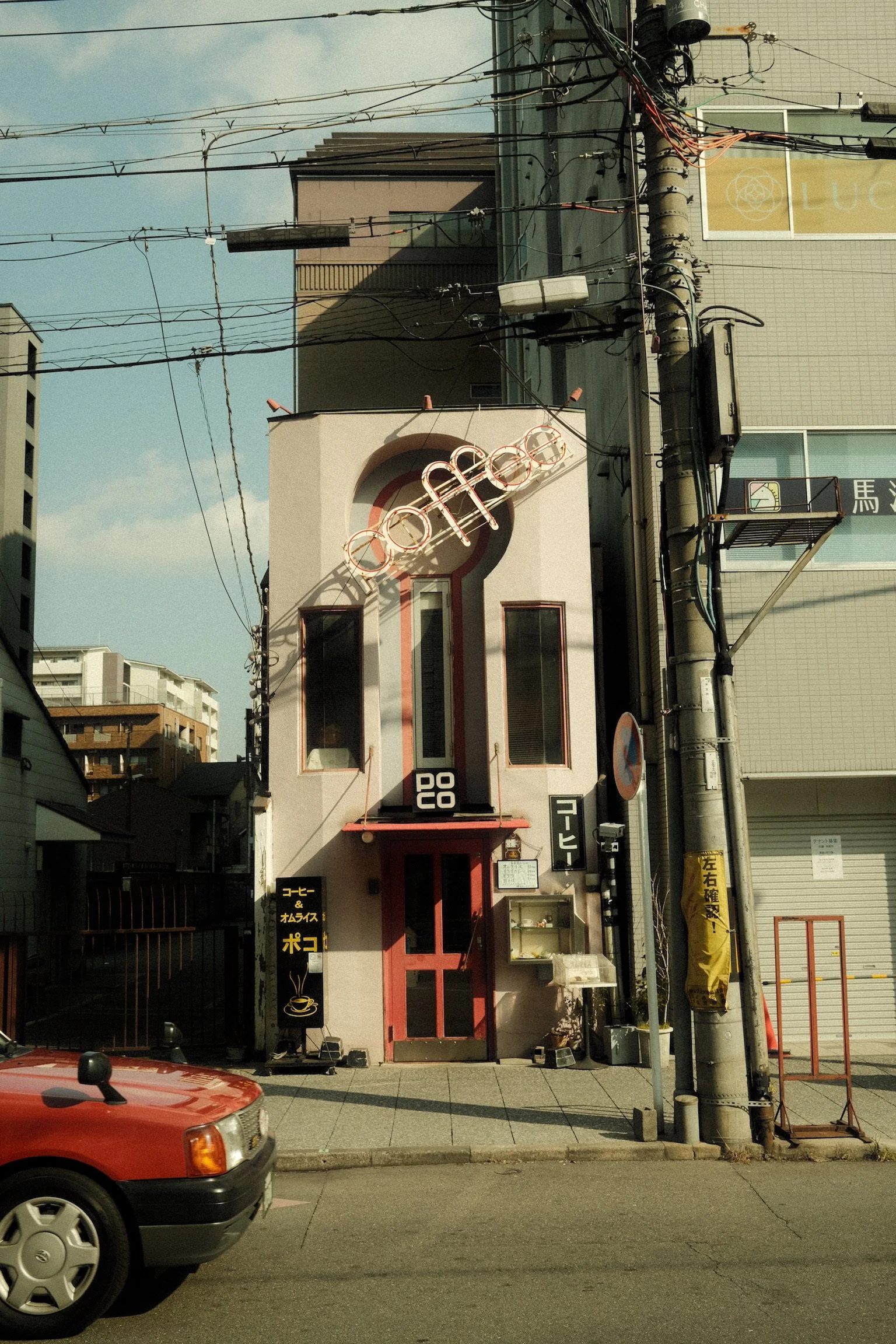A small building with a sign that has the word 'coffee' in English, Japanese characters, and a drawing of a steaming coffee cup. There is a red door, and the building is white with pink accents. A blackboard sign is outside, and a utility pole with w