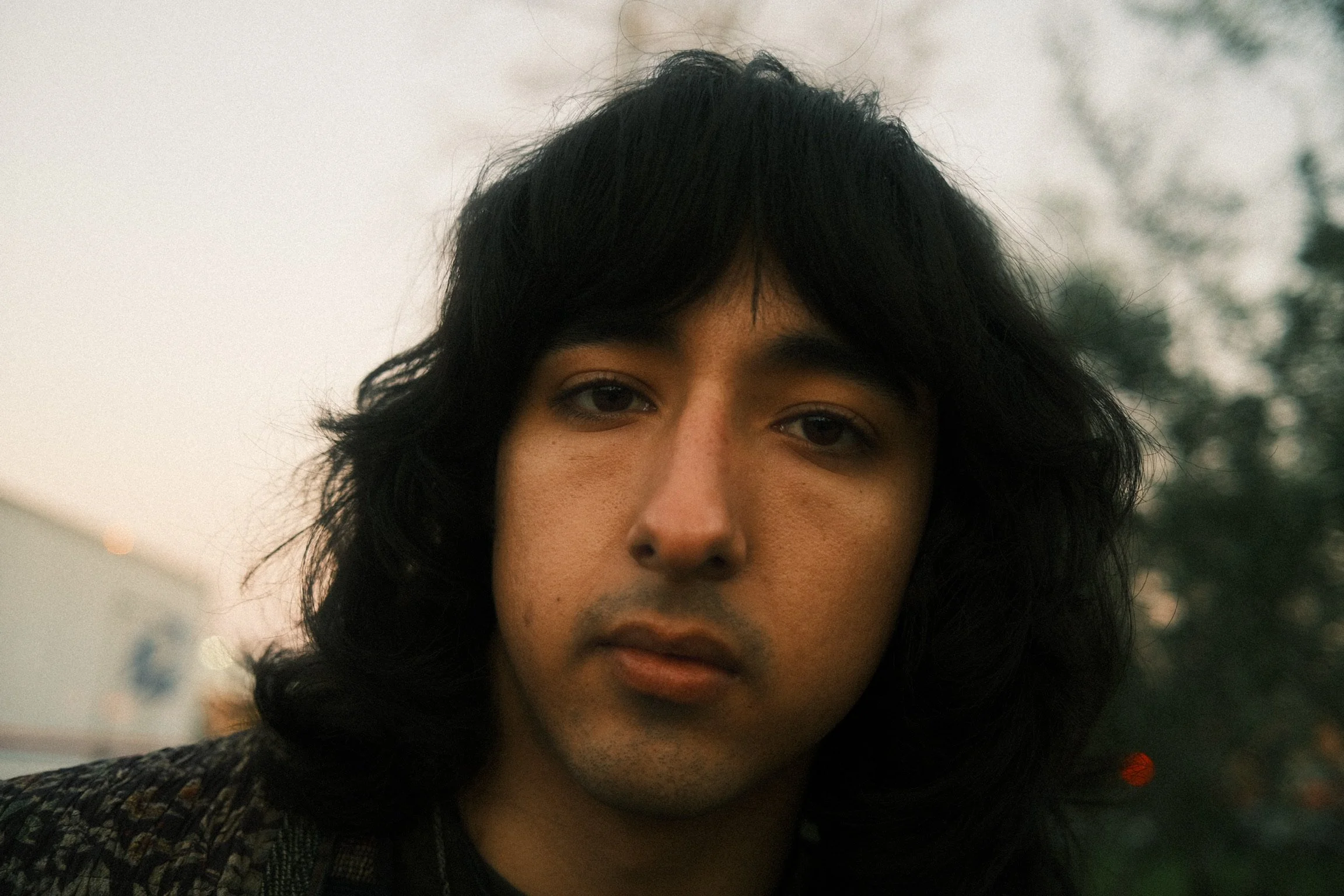Close-up of a person's face with dark hair, brown eyes, and slightly tanned skin, outdoors with blurred background.