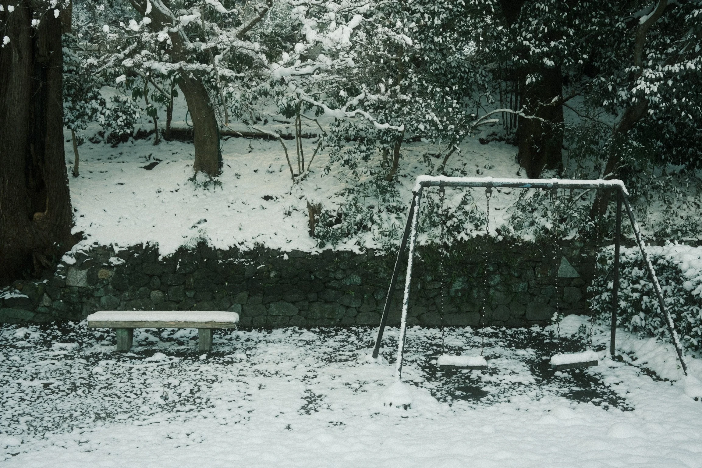 Snow-covered playground with a swing set, a park bench, and trees in the background.