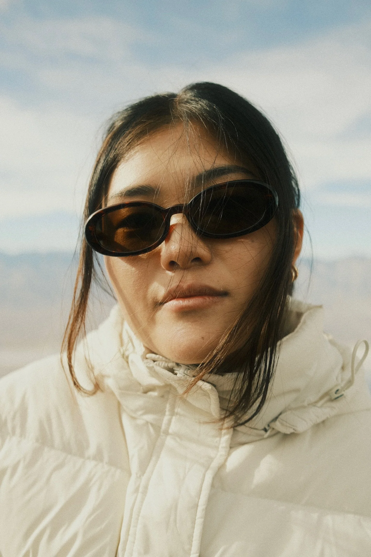 A close-up of a woman wearing large black sunglasses and a white puffer jacket outdoors against a cloudy sky.