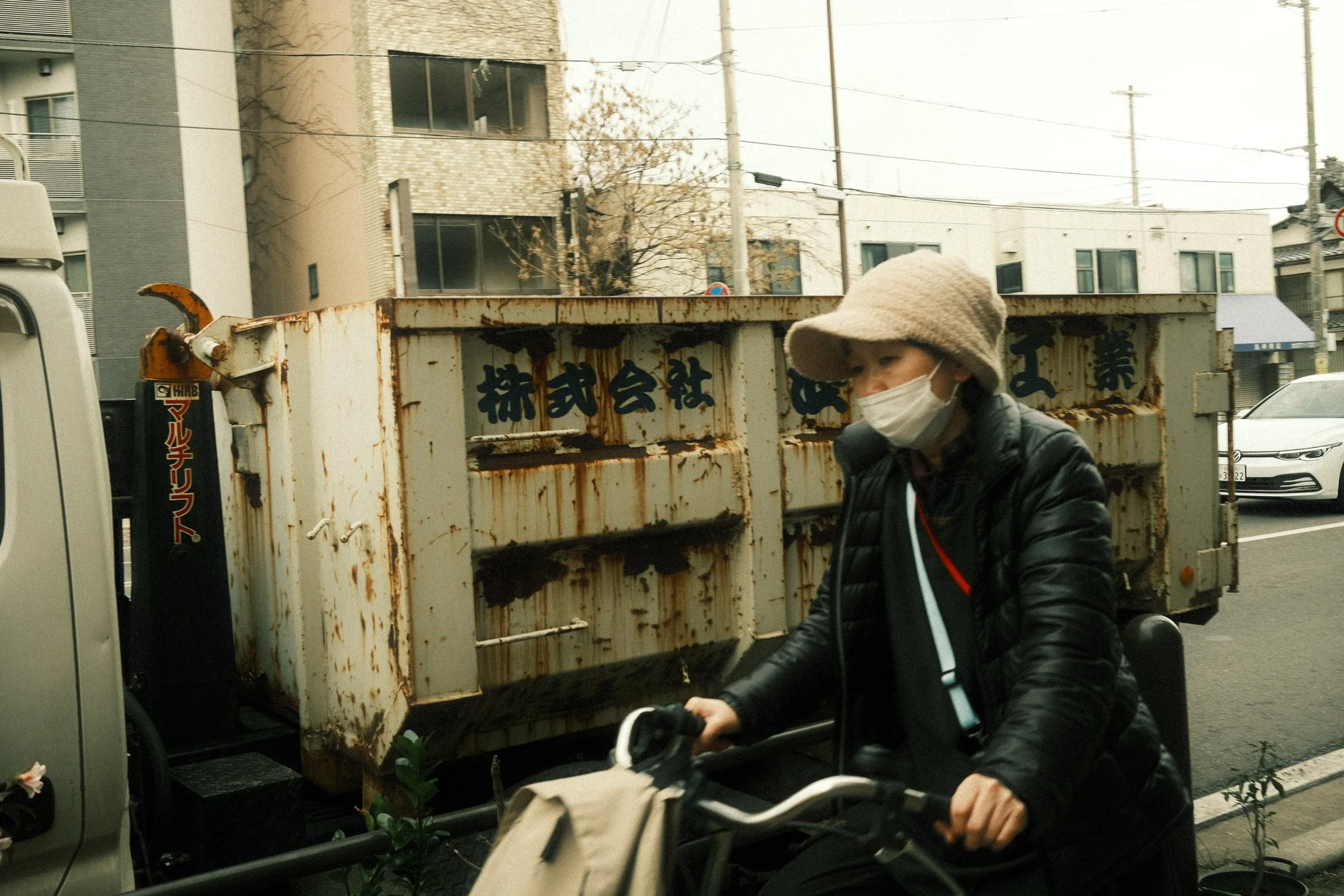 Person wearing a face mask and a beige hat riding a bicycle past a rusty, old truck with Japanese writing on it in an urban setting.