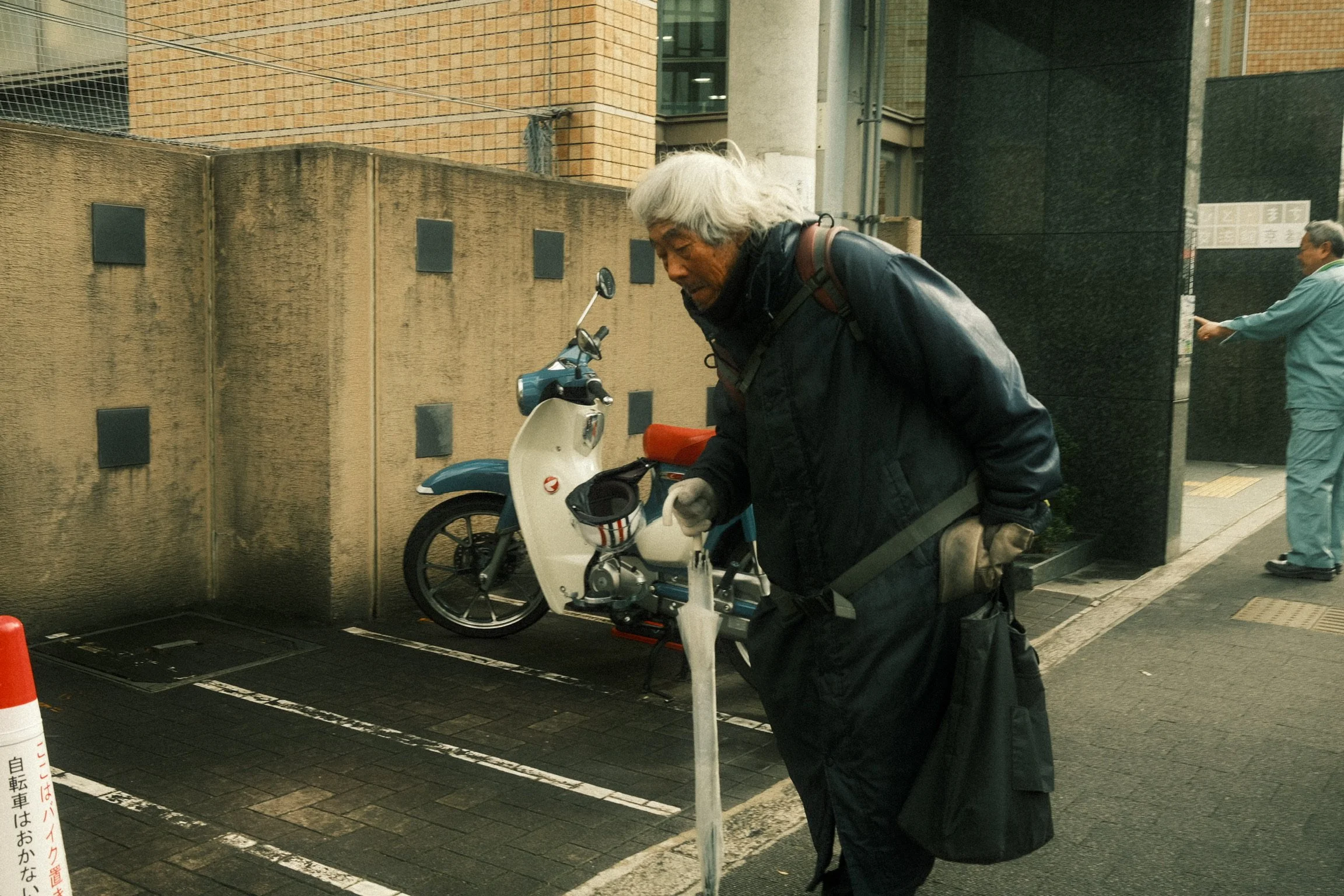 An elderly man with gray hair loading a scooter at a parking lot near a sidewalk, with two other men in the background, one reading a sign and the other walking.