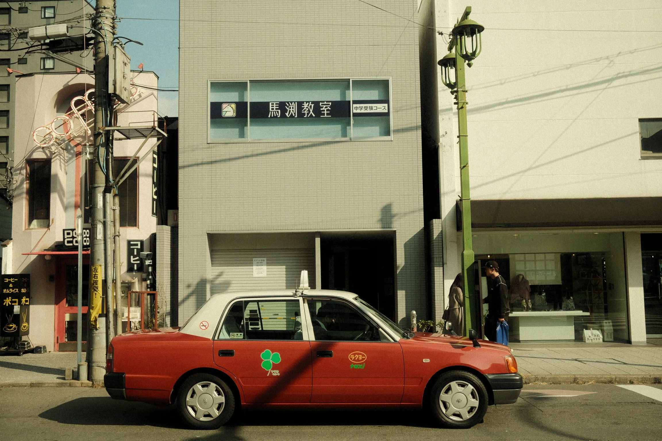 A red compact car with green four-leaf clover decal and taxi signs parked on a city street in front of a modern building with Japanese signage and people walking by.