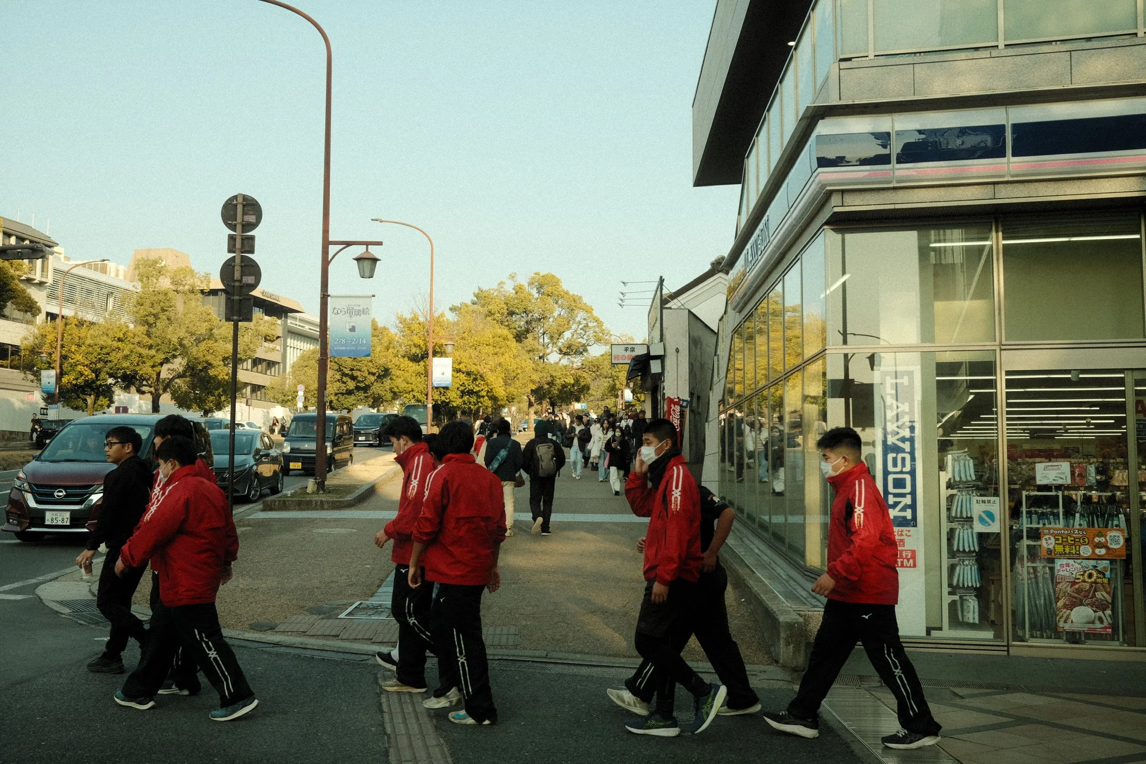 Street scene in Japan with a group of young people wearing red jackets and black pants crossing the sidewalk. There are several cars on the street, a building with glass windows to the right, and trees in the background. People are wearing masks, and